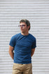 Young man with glasses wearing a blue t-shirt and beige pants standing against white brick wall