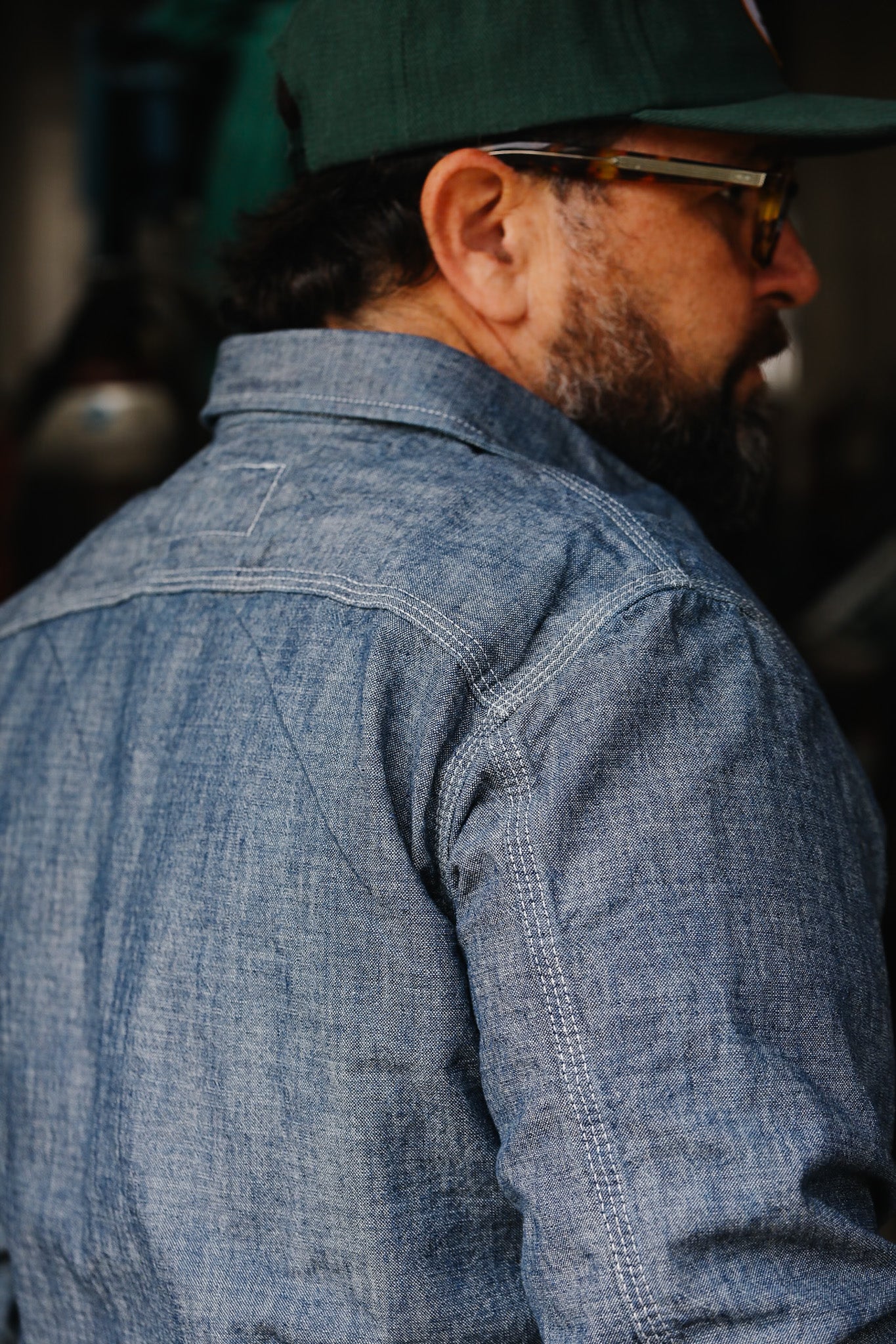 Man wearing chambray work shirt and green baseball cap, side profile in workshop setting