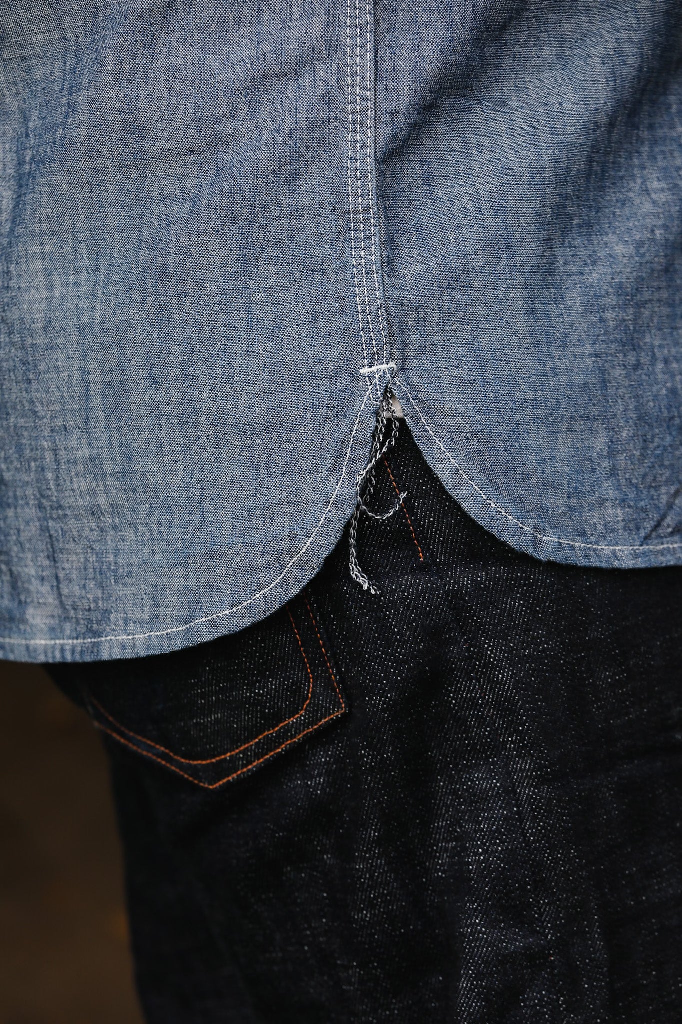 Close-up of chambray shirt hem with raw edges over dark denim jeans pocket with orange stitching