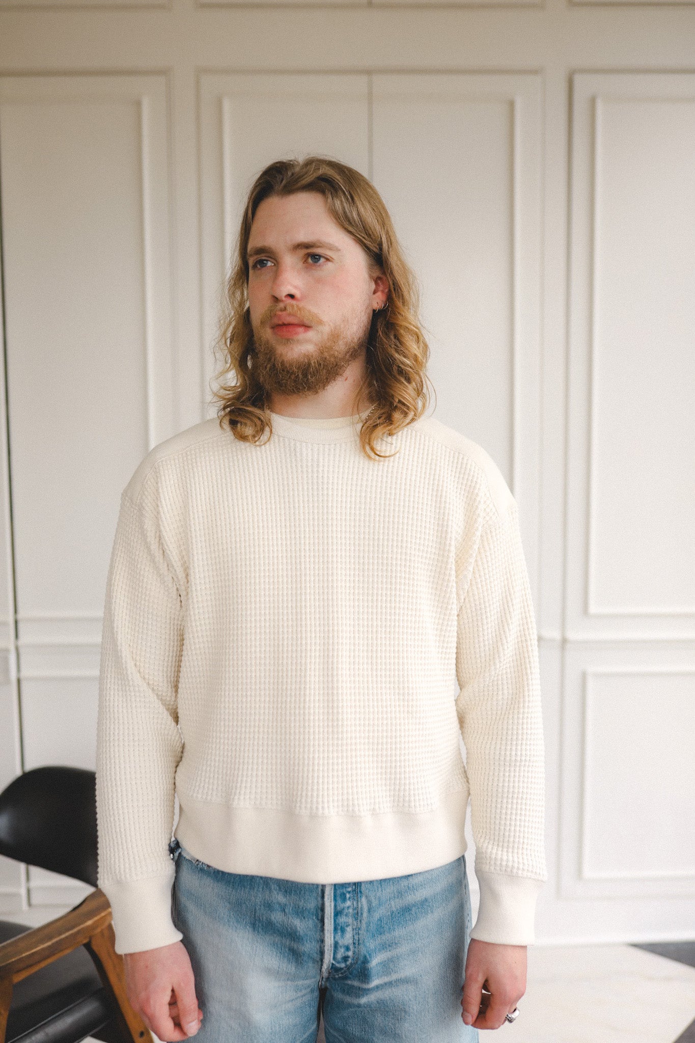Man wearing a cream sweater and blue jeans standing against a white paneled wall.