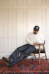 Man sitting on a stool wearing a white sweater and dark jeans, with a cap on his head, against a paneled wall.