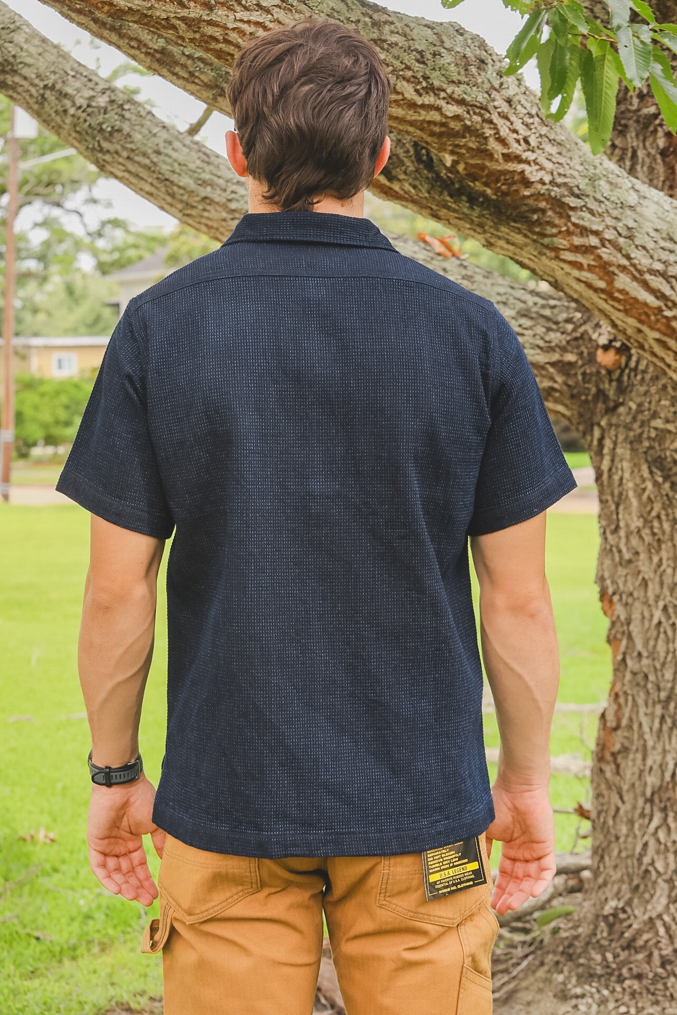 Person wearing a navy blue shirt and brown pants standing next to a tree