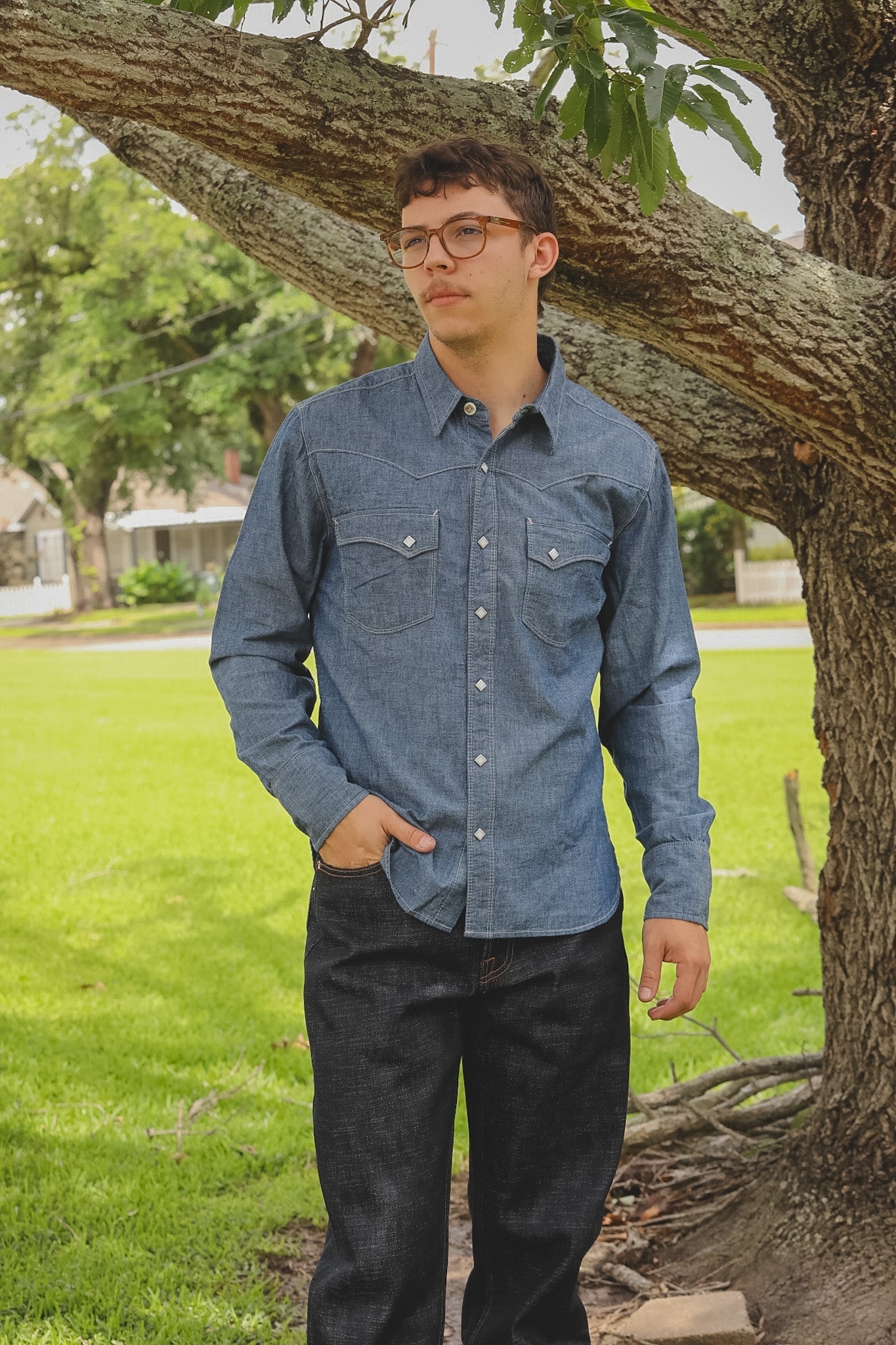 Man wearing a blue denim shirt standing under a tree in a park.
