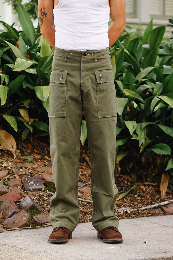 Person wearing green cargo pants and a white tank top standing outdoors with plants in the background.
