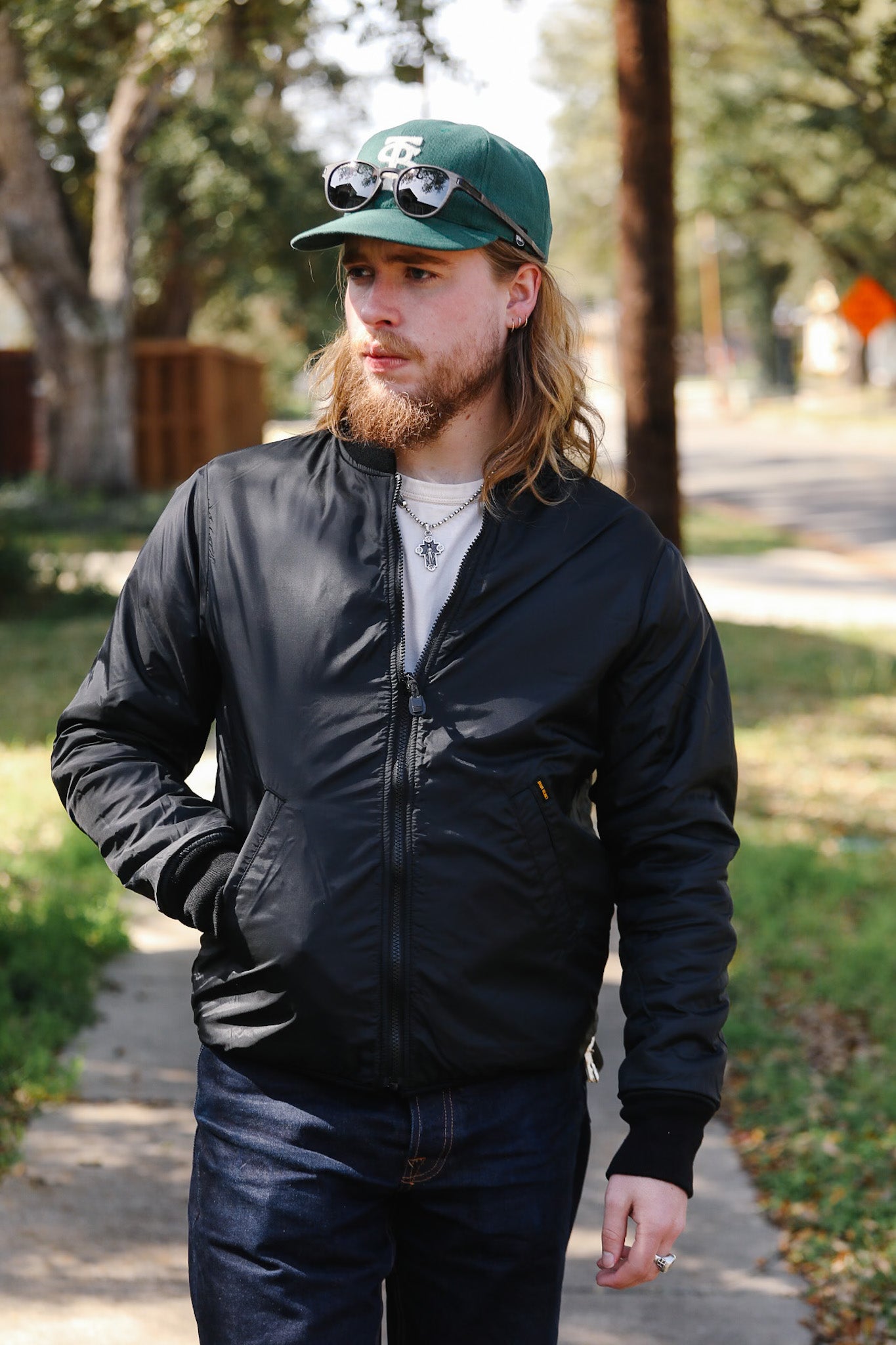 Man wearing a black bomber jacket and green cap with sunglasses on top, standing outdoors.