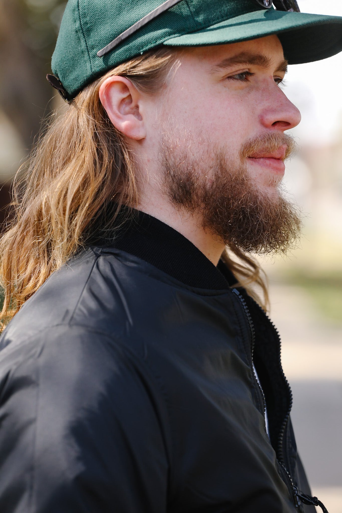 Man with long hair and beard wearing a green cap and black jacket outdoors