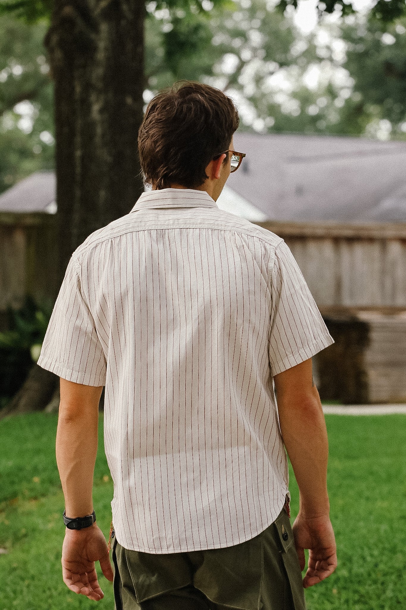 Man wearing a light-colored striped shirt and sunglasses, standing in a grassy area with trees and a house in the background.