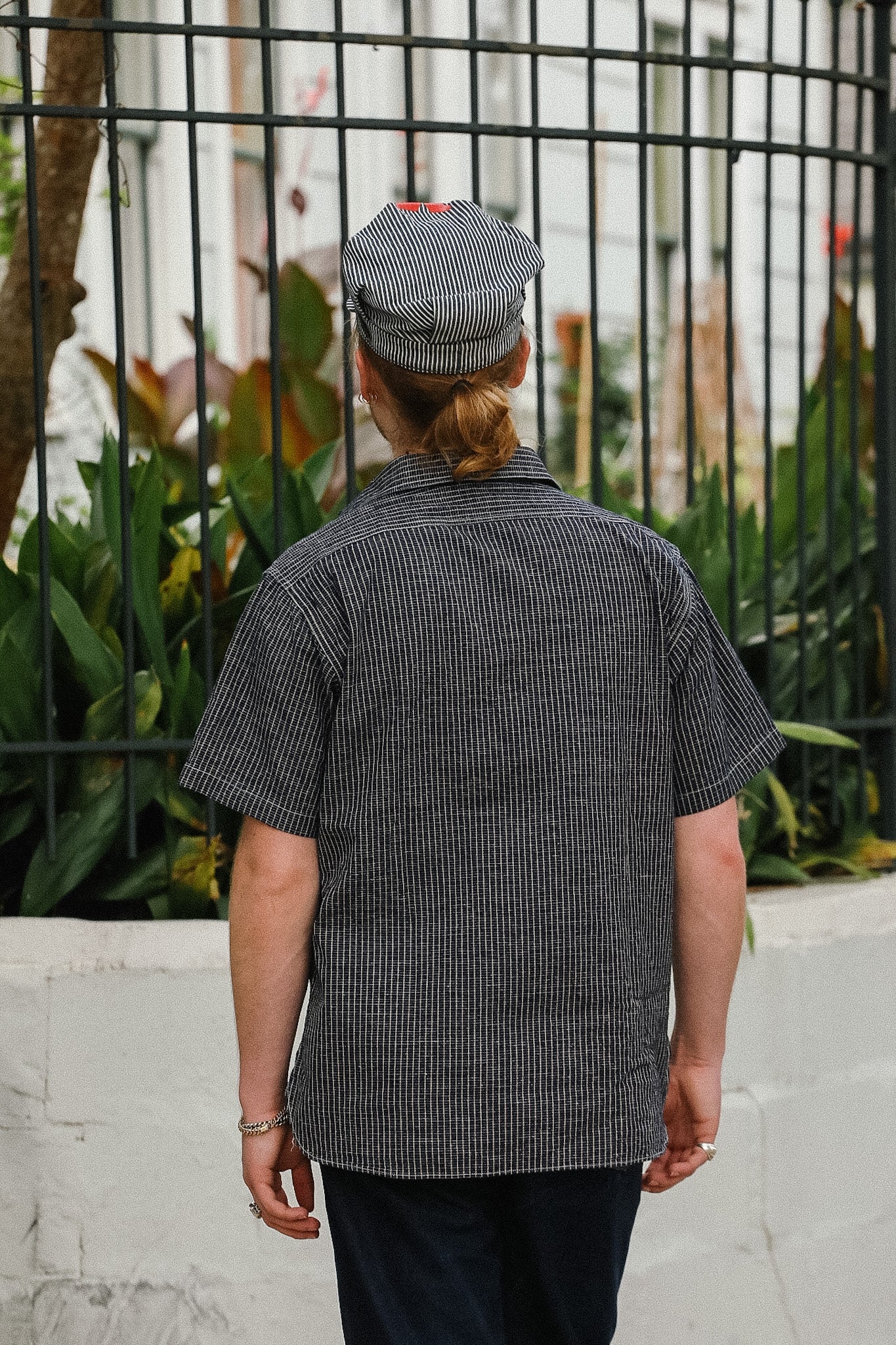 Person wearing a black and white checkered shirt with a cap, standing in front of a metal fence and plants.