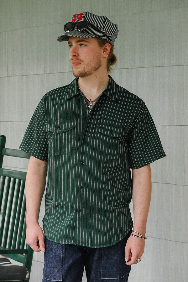 Man wearing a green striped shirt and cap standing against a tiled wall.