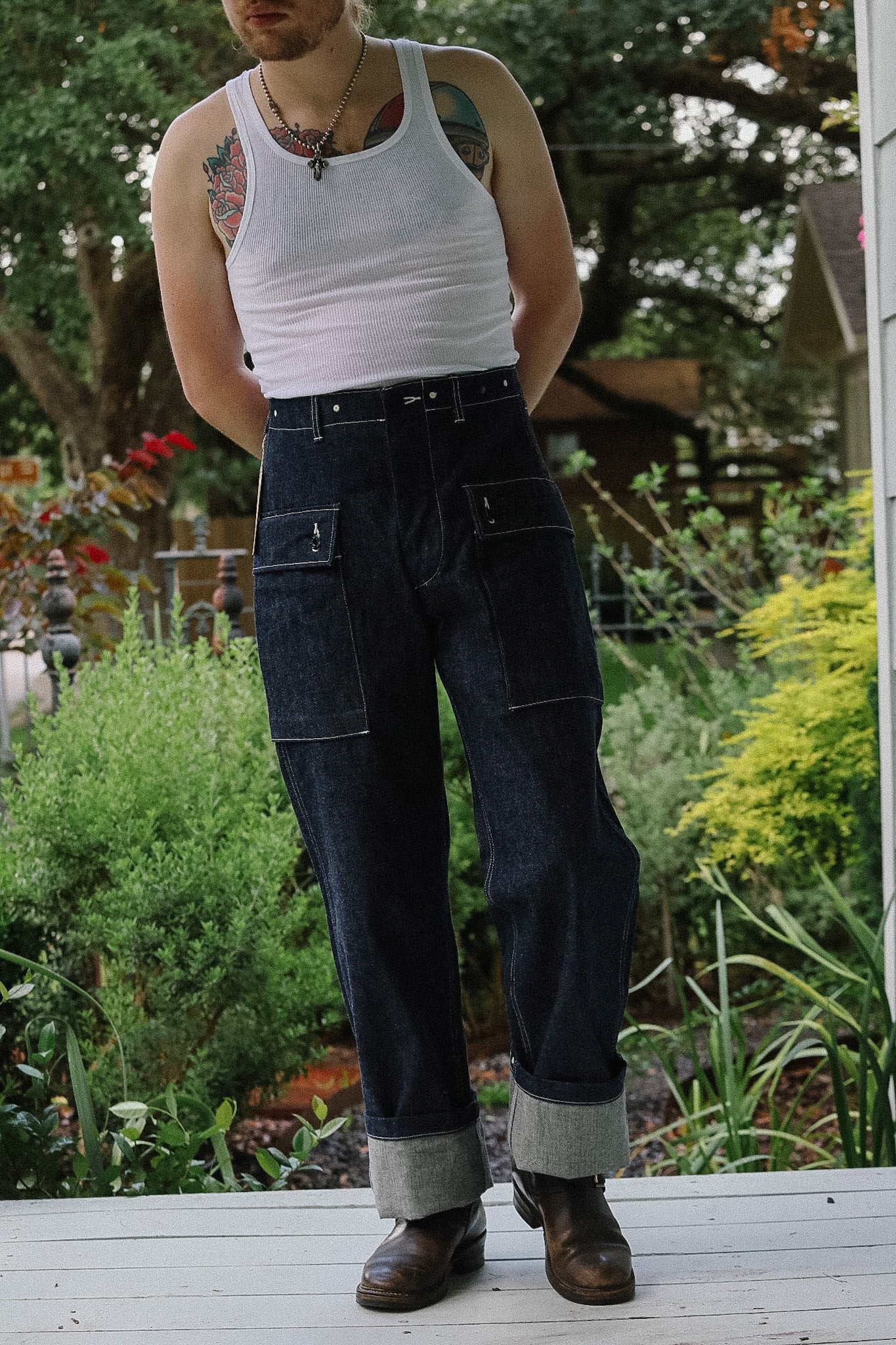 Person wearing a white tank top and dark jeans standing on a wooden deck with a garden background.