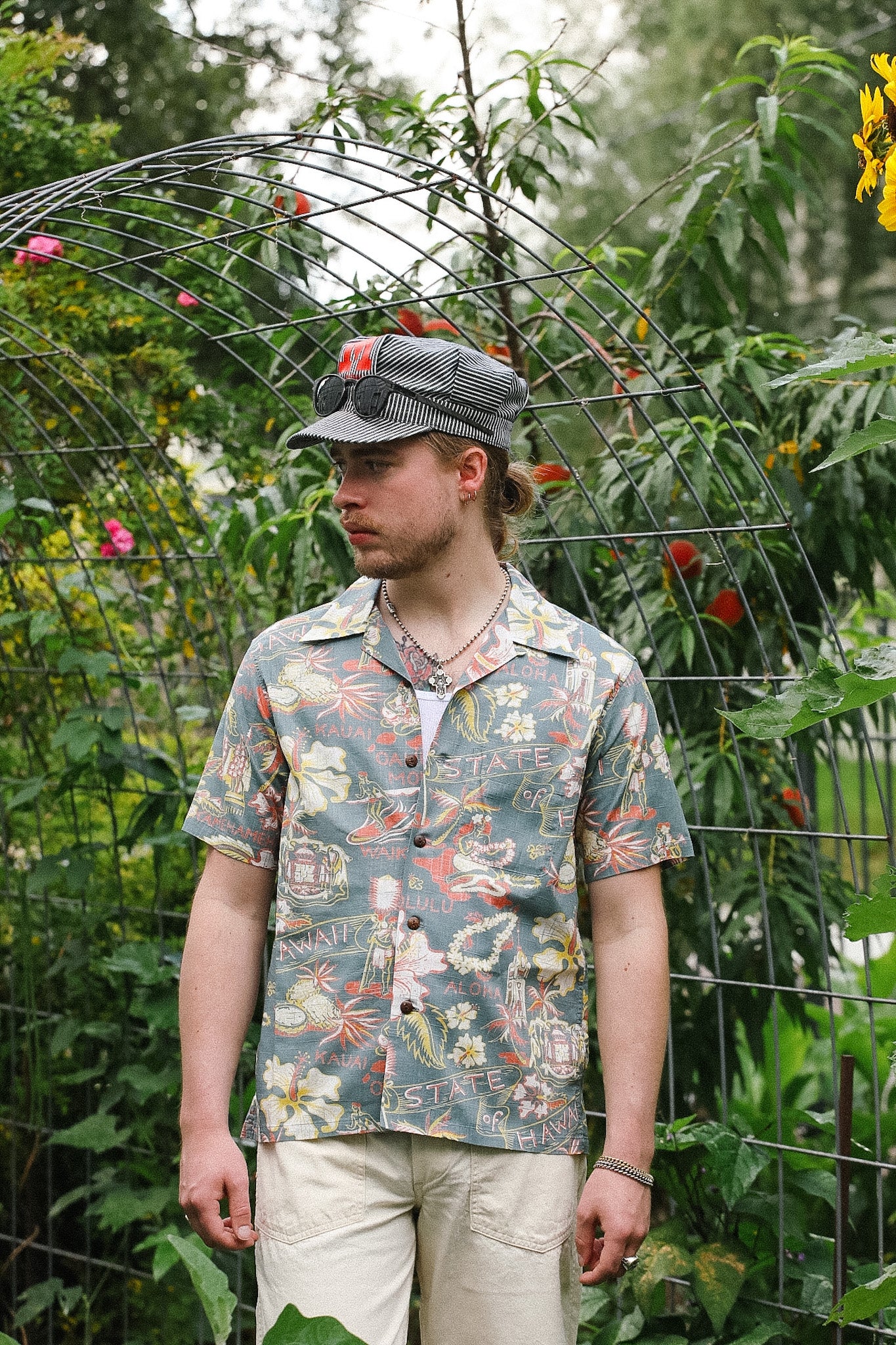 Man wearing a patterned shirt and cap standing in a garden with greenery and flowers.