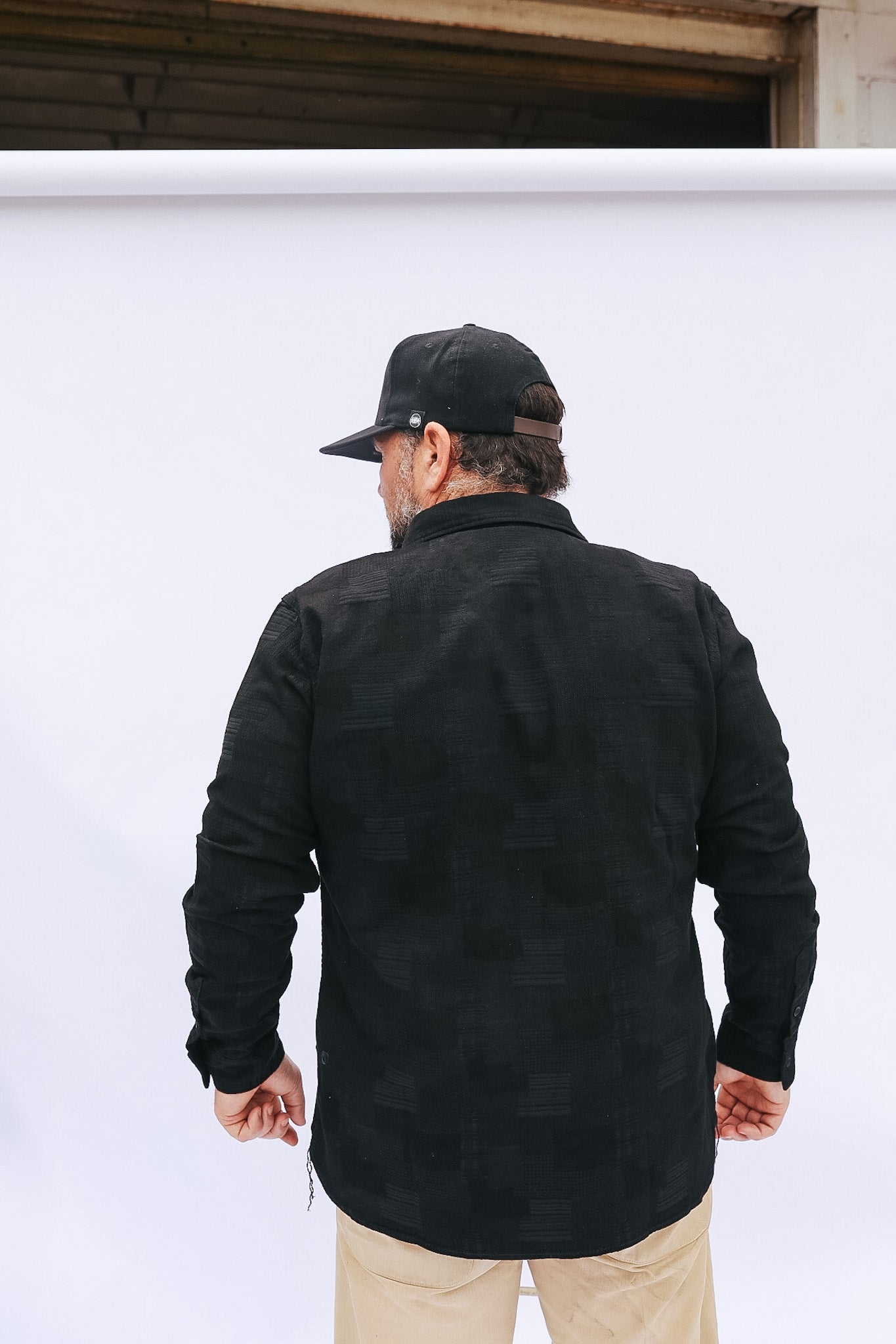 Man wearing a black jacket and cap standing against a white wall.