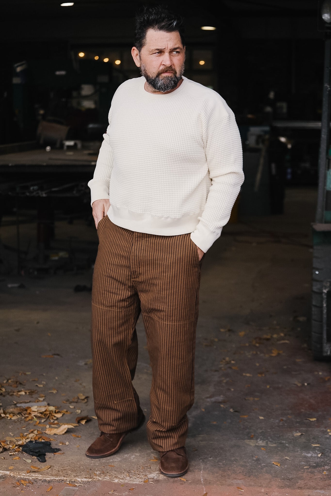Man wearing a white sweater and brown checkered pants standing in a dimly lit room.