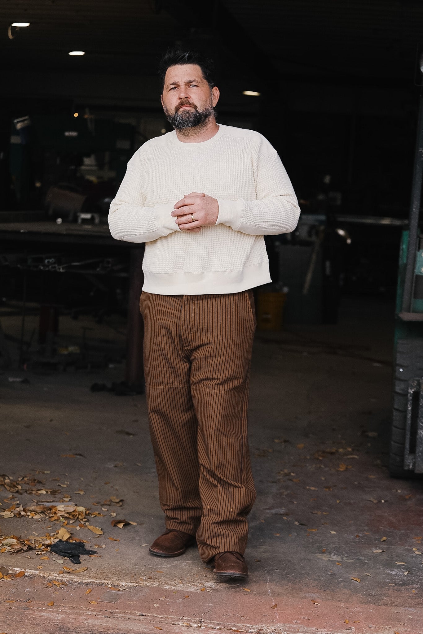 Man wearing a white sweater and brown pants standing in an indoor setting with dark background