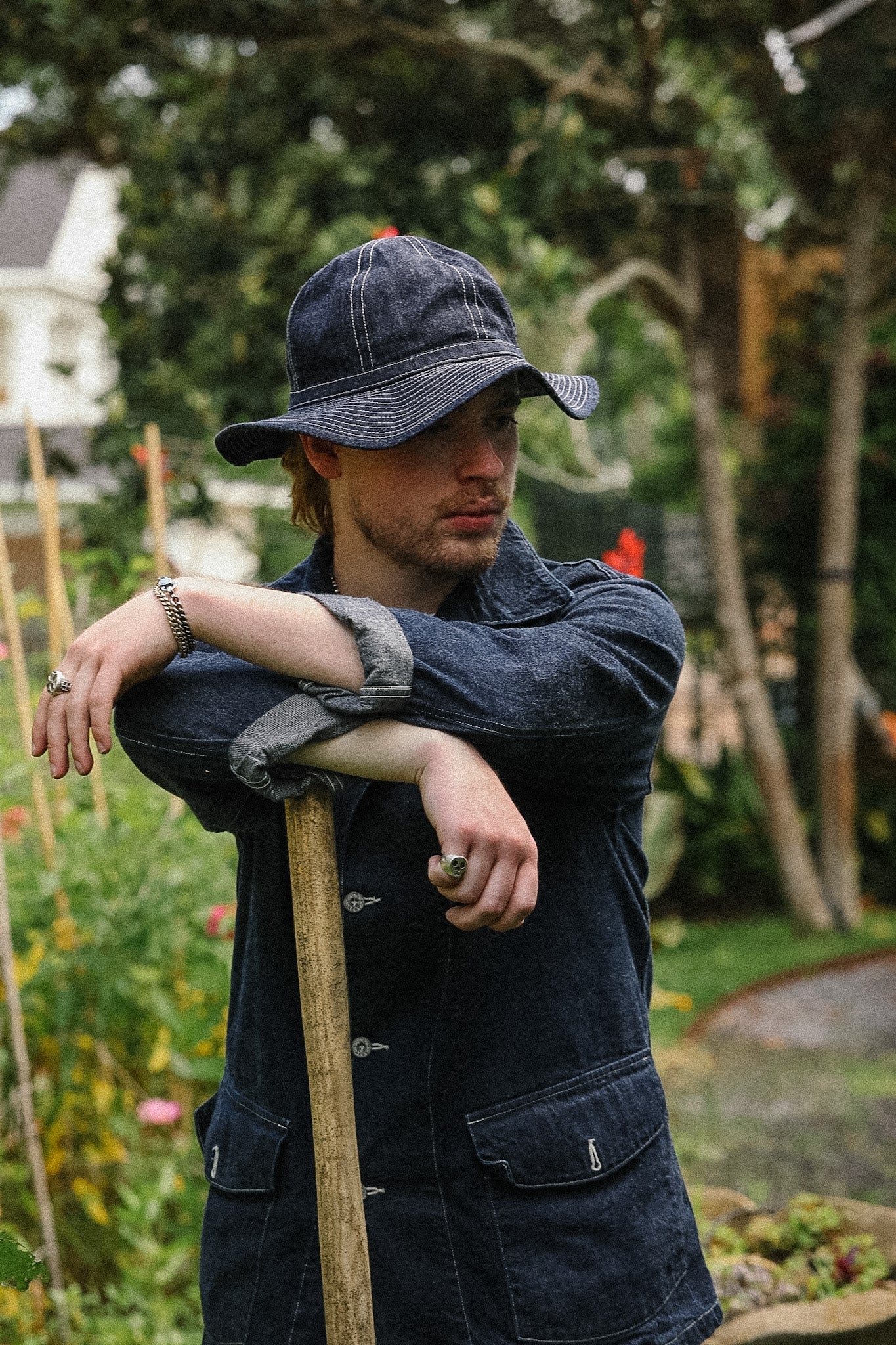 Man in a garden wearing a denim jacket and cap, holding a shovel.