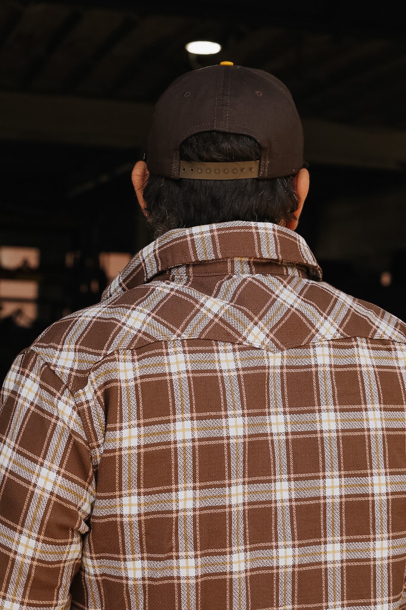 Person wearing a brown plaid shirt and cap from behind, in a dark indoor setting.