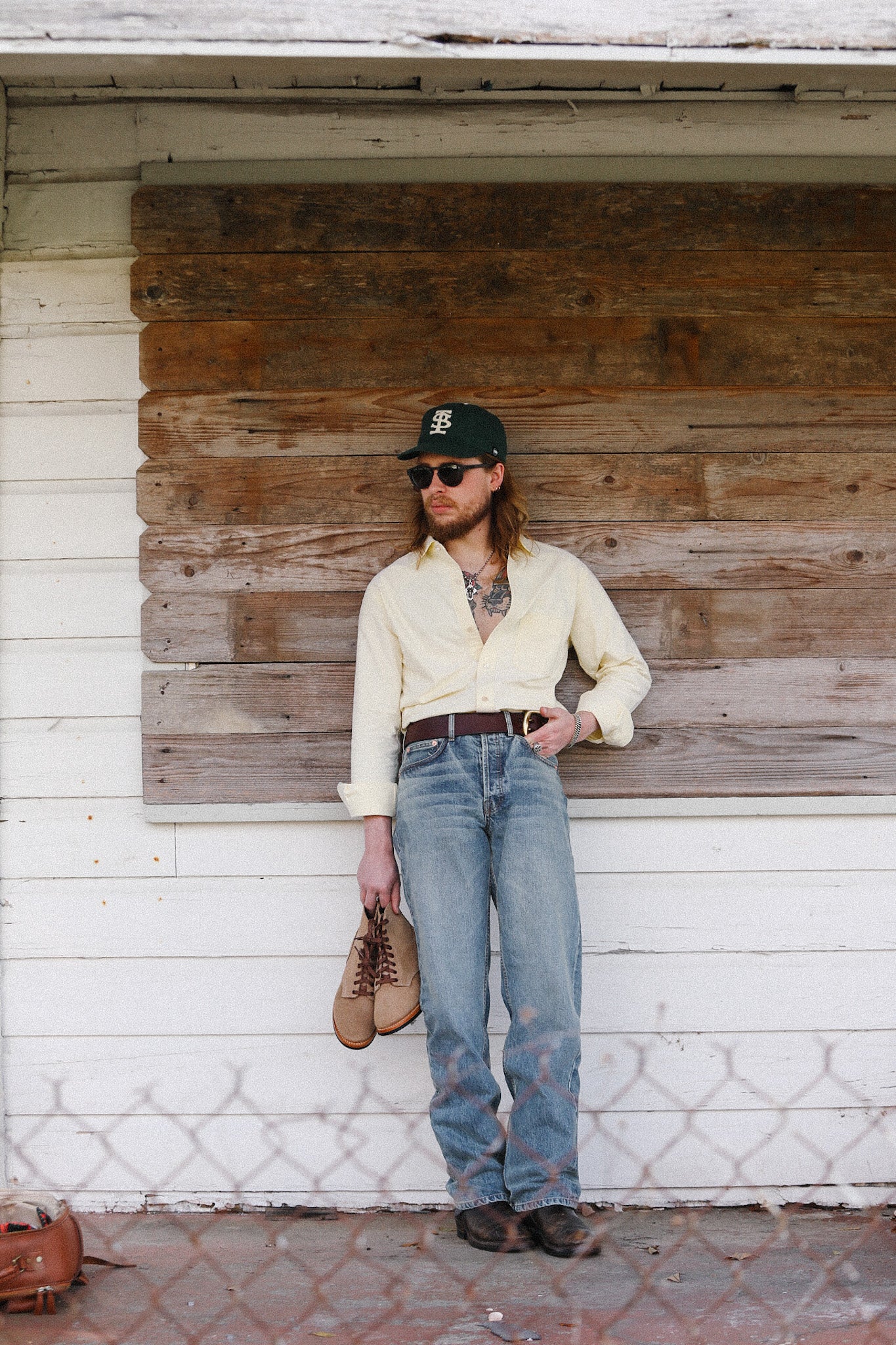 Person wearing a white shirt, blue jeans, and a cap leaning against a rustic wooden wall.