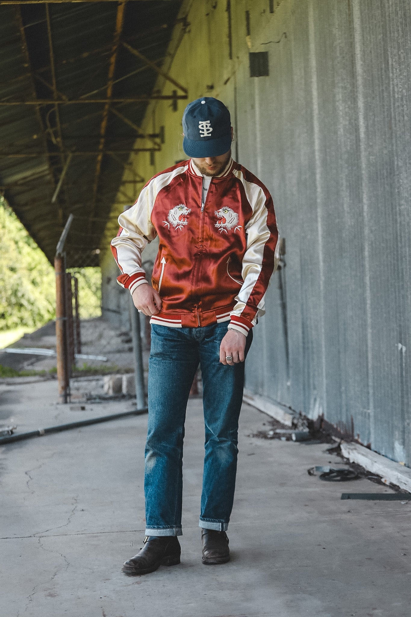 A man wearing a red and cream satin bomber jacket with embroidered dragon designs on the chest, blue jeans, and a navy baseball cap, standing in an industrial setting.
