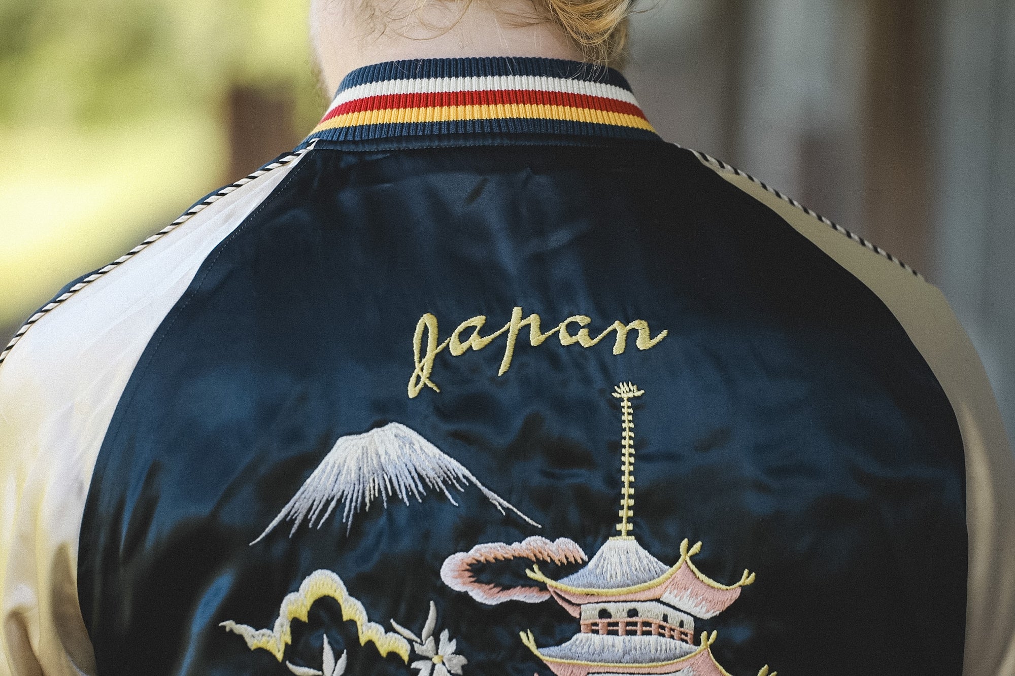 Close-up back view of a person wearing a satin bomber jacket with cream sleeves, a navy collar with red and yellow stripes, and gold embroidered Japanese motifs including Mount Fuji, a pagoda, clouds, and the word 'Japan'.