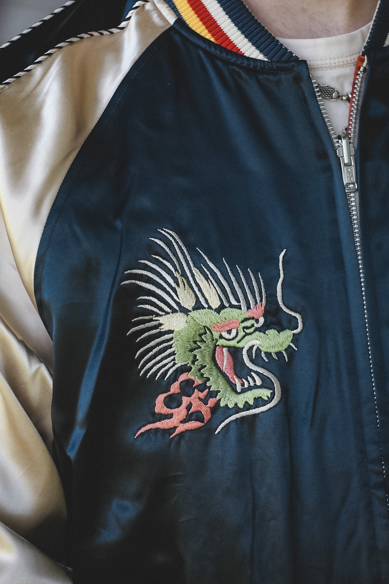 Close-up of a satin bomber jacket worn by a person, featuring a green and pink embroidered dragon motif on the chest, cream-colored satin sleeves, and a striped collar.