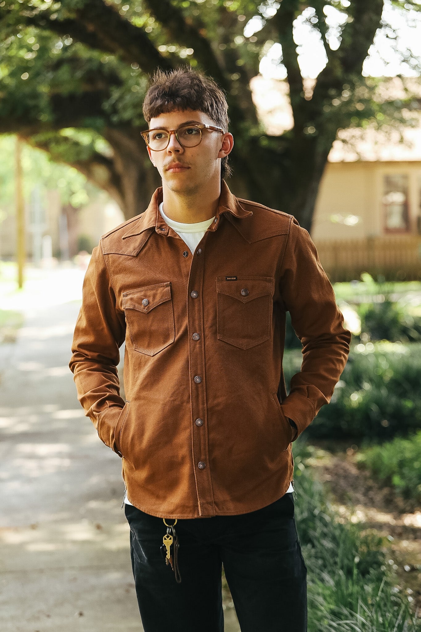 A man wearing a brown corduroy western-style button-up shirt and black pants, standing outdoors on a path with trees in the background.