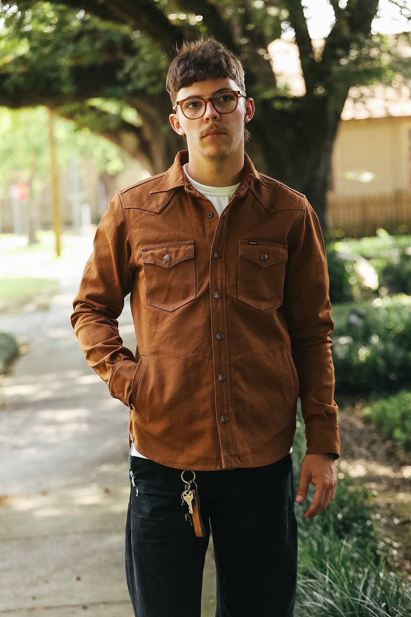 A man wearing a brown corduroy western-style button-up shirt with chest pockets, paired with black jeans and glasses, standing outdoors on a paved path with trees in the background.