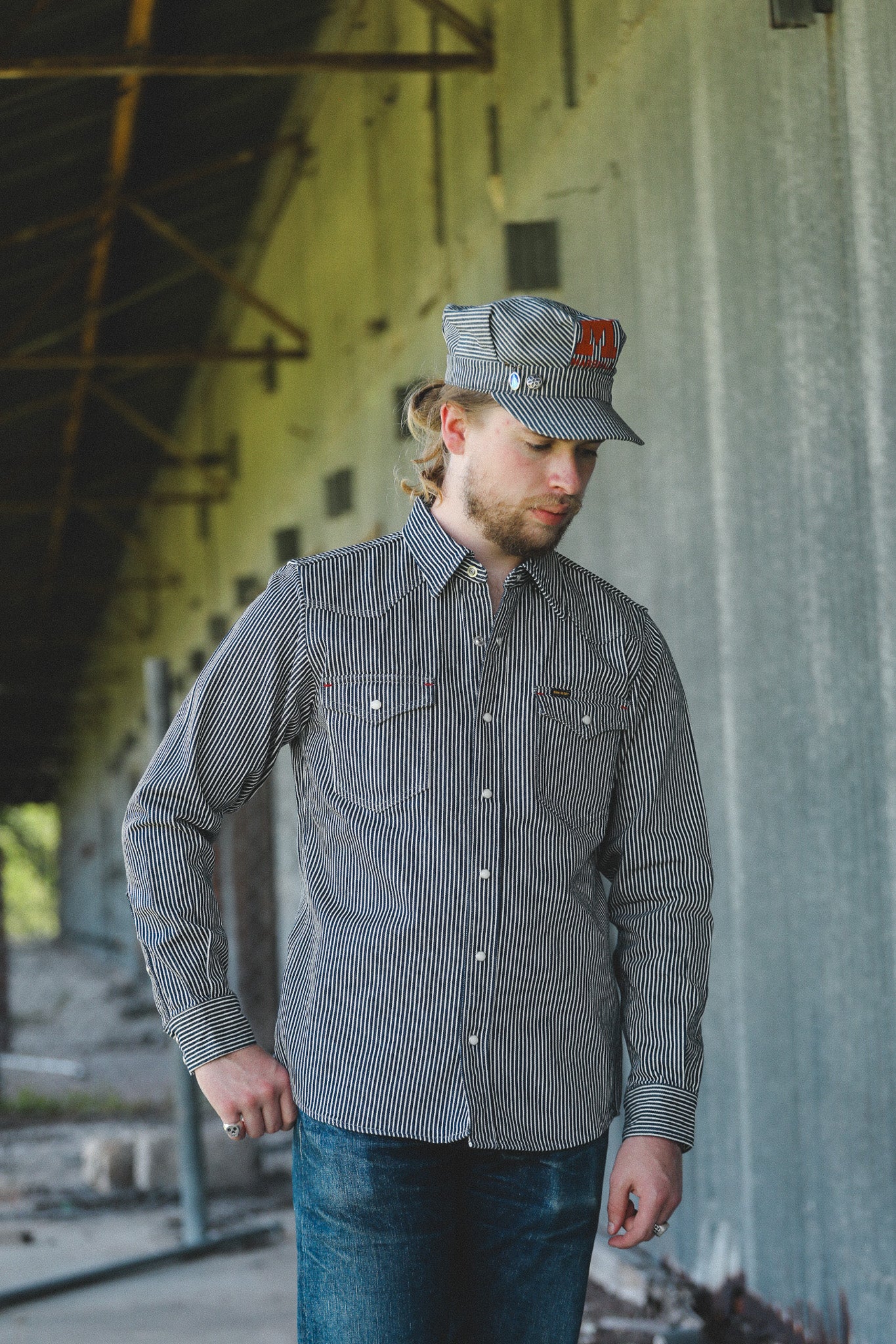 A man wearing a gray and black vertically striped button-down shirt and a matching gray cap with a red logo, standing in an industrial warehouse setting.