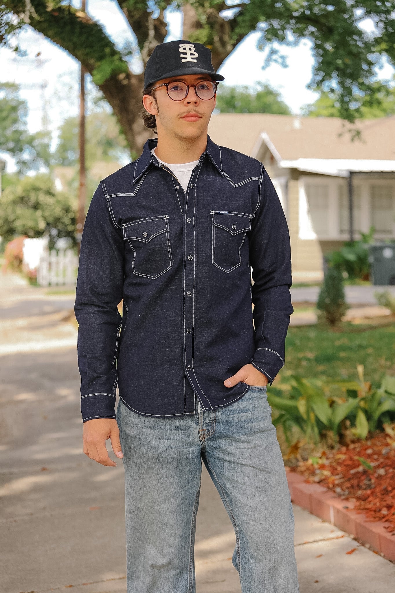 Man wearing a dark denim shirt and cap standing on a sidewalk with trees and houses in the background.