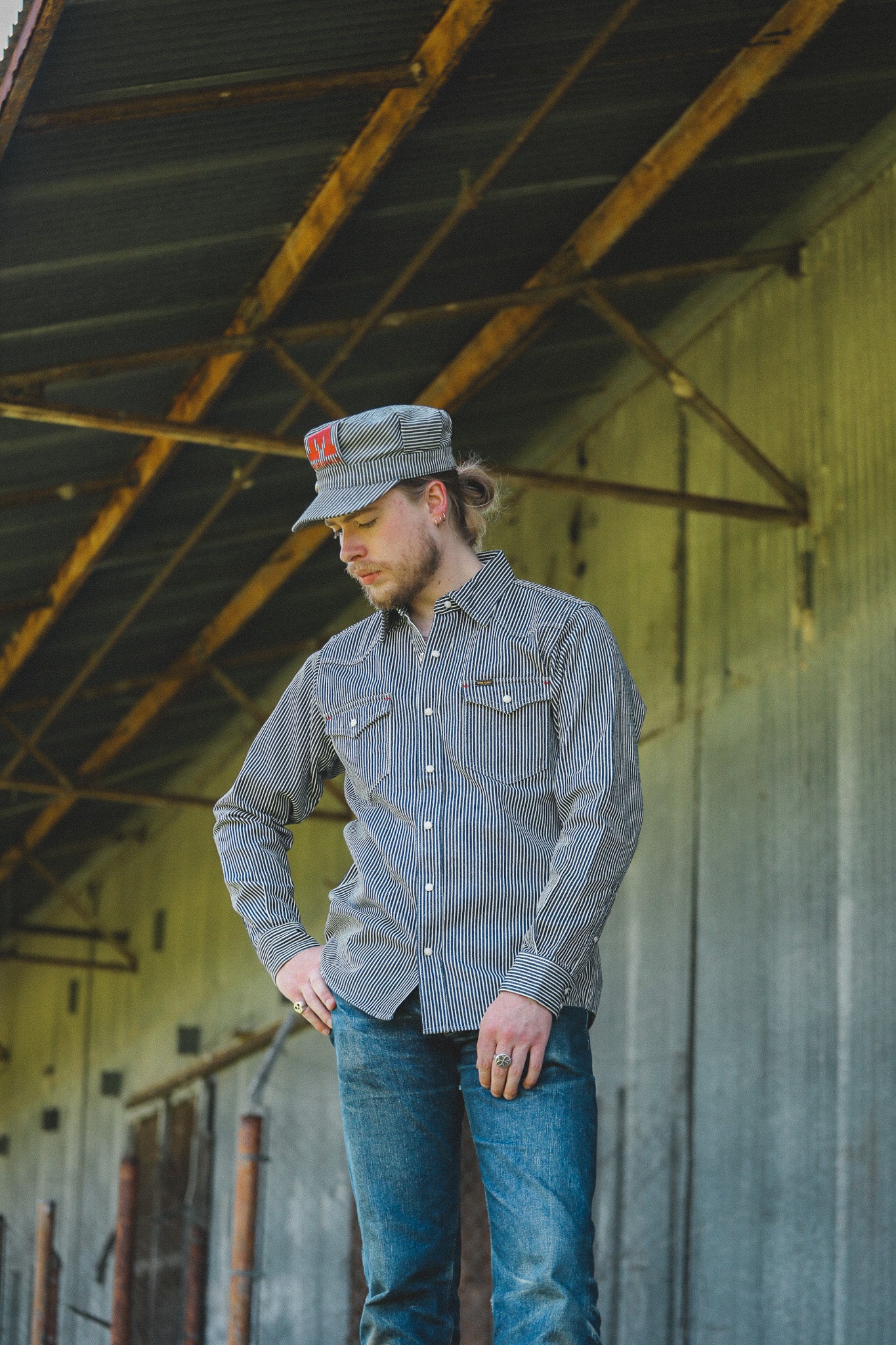 A man wearing a gray and white striped long-sleeve button-down shirt, blue jeans, and a matching gray cap with a red logo, standing in an industrial warehouse setting.