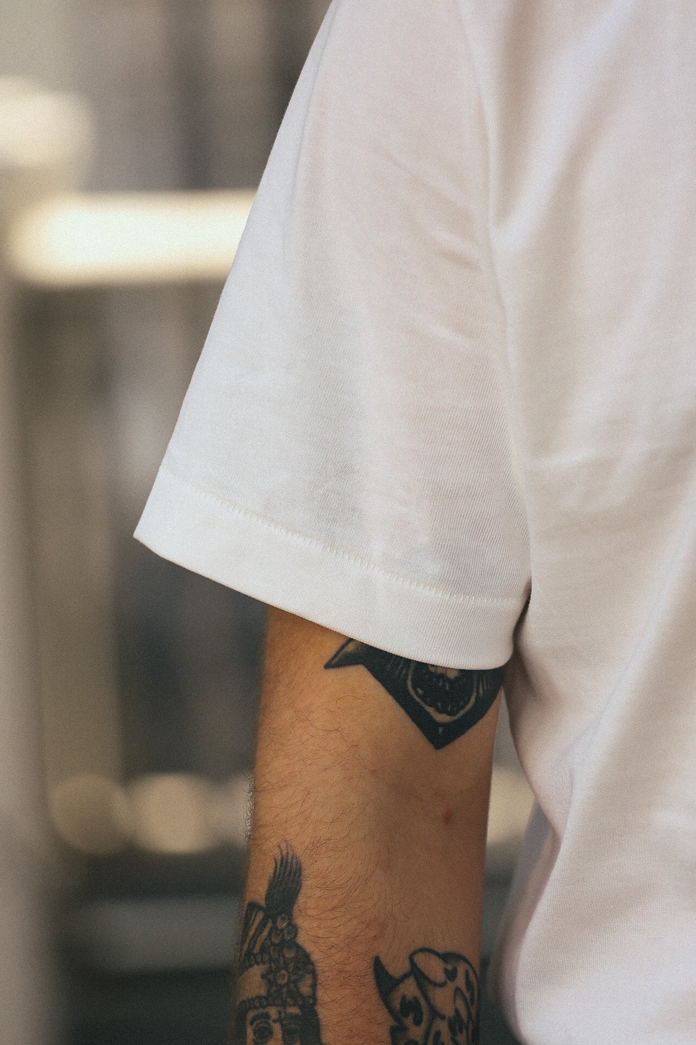 Close-up of a person wearing a white short-sleeve t-shirt, showing the sleeve and upper arm with visible black tattoos against a blurred indoor background.