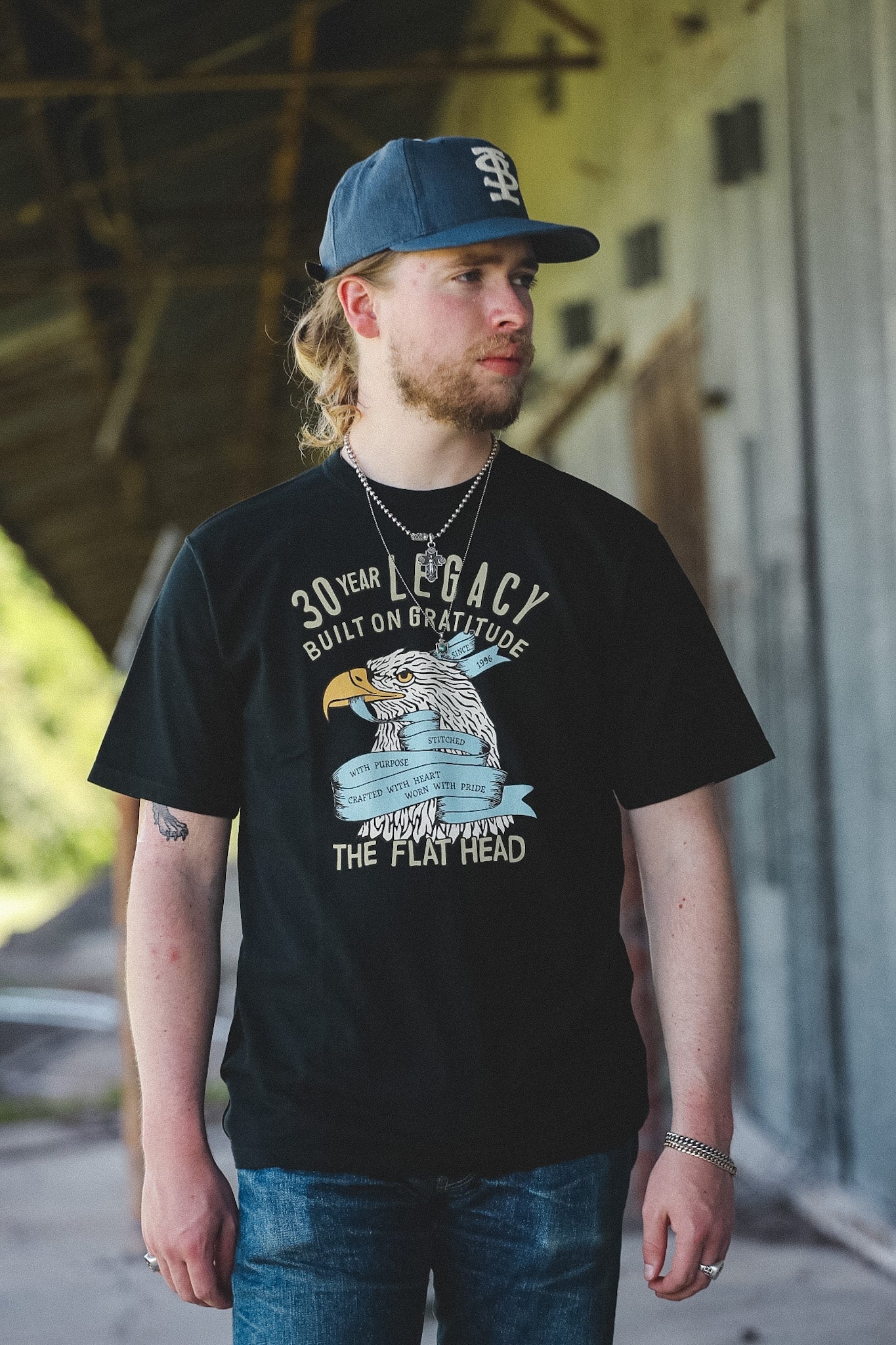 A man wearing a black graphic t-shirt with a distressed eagle design and text, a blue baseball cap, and blue jeans, standing in front of a rustic wooden structure.