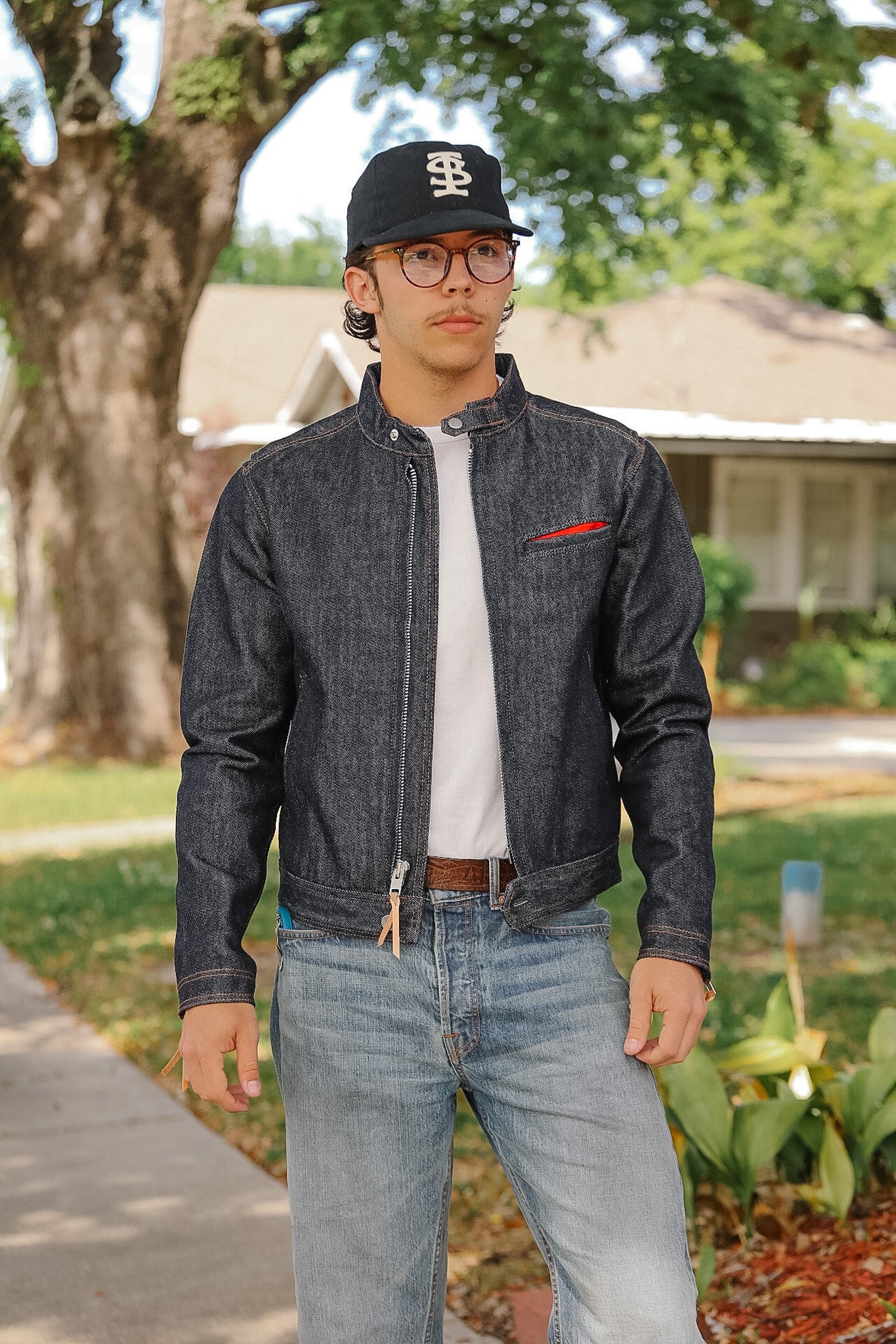 Man wearing a denim jacket and cap standing outdoors with trees and houses in the background