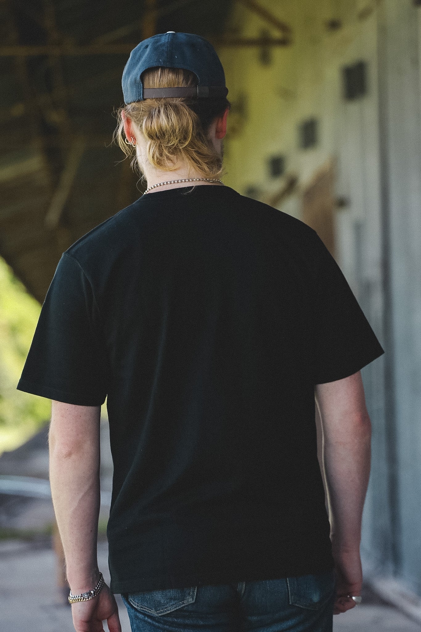 A person viewed from behind wearing a black short-sleeve t-shirt, blue jeans, a blue baseball cap, and a silver chain necklace, standing in a rustic covered walkway.