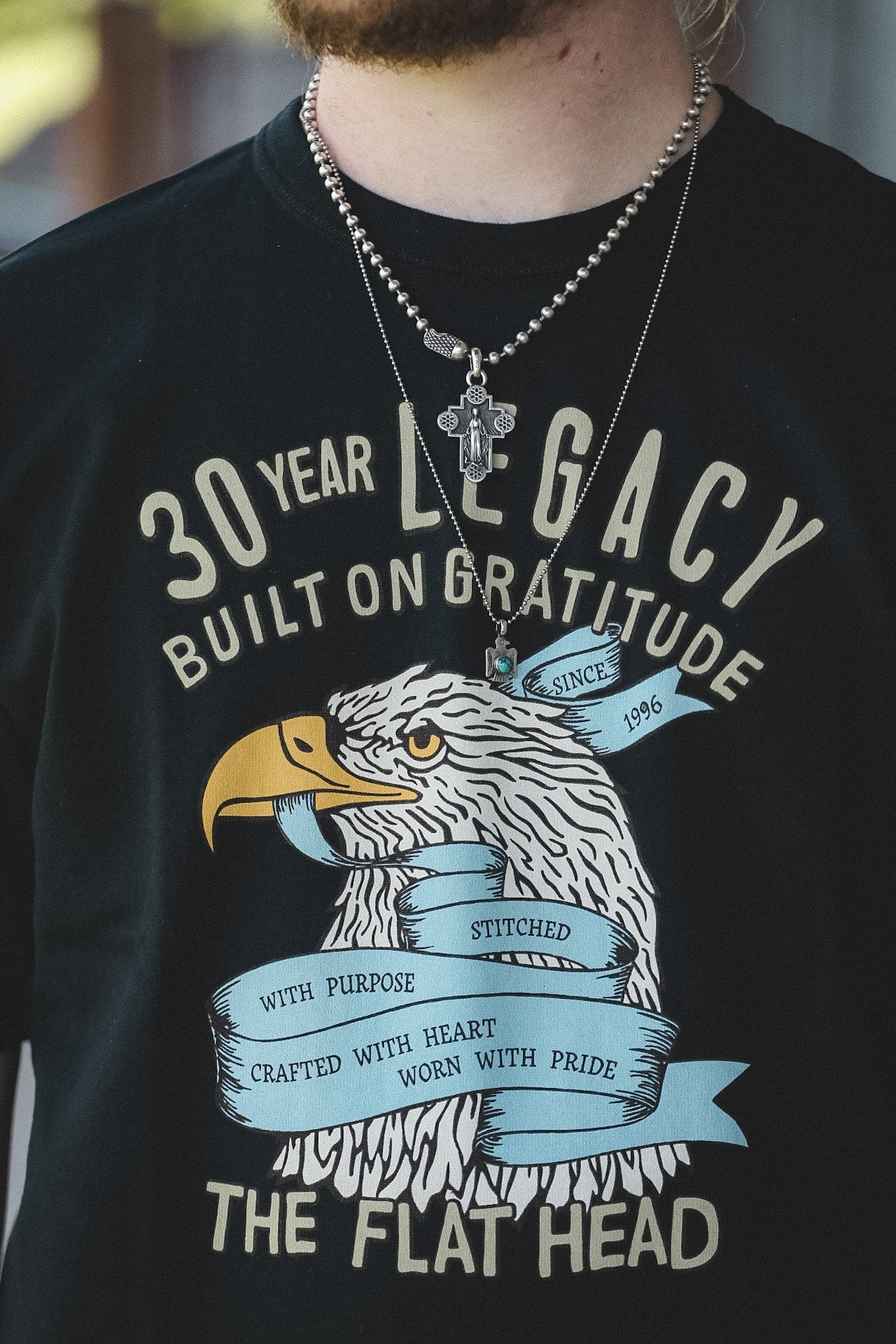 A close-up of a person wearing a black graphic t-shirt with a vintage-style eagle design and cream-colored text reading '30 YEAR LEGACY BUILT ON GRATITUDE' and 'THE FLAT HEAD'. The person is also wearing layered silver necklaces.