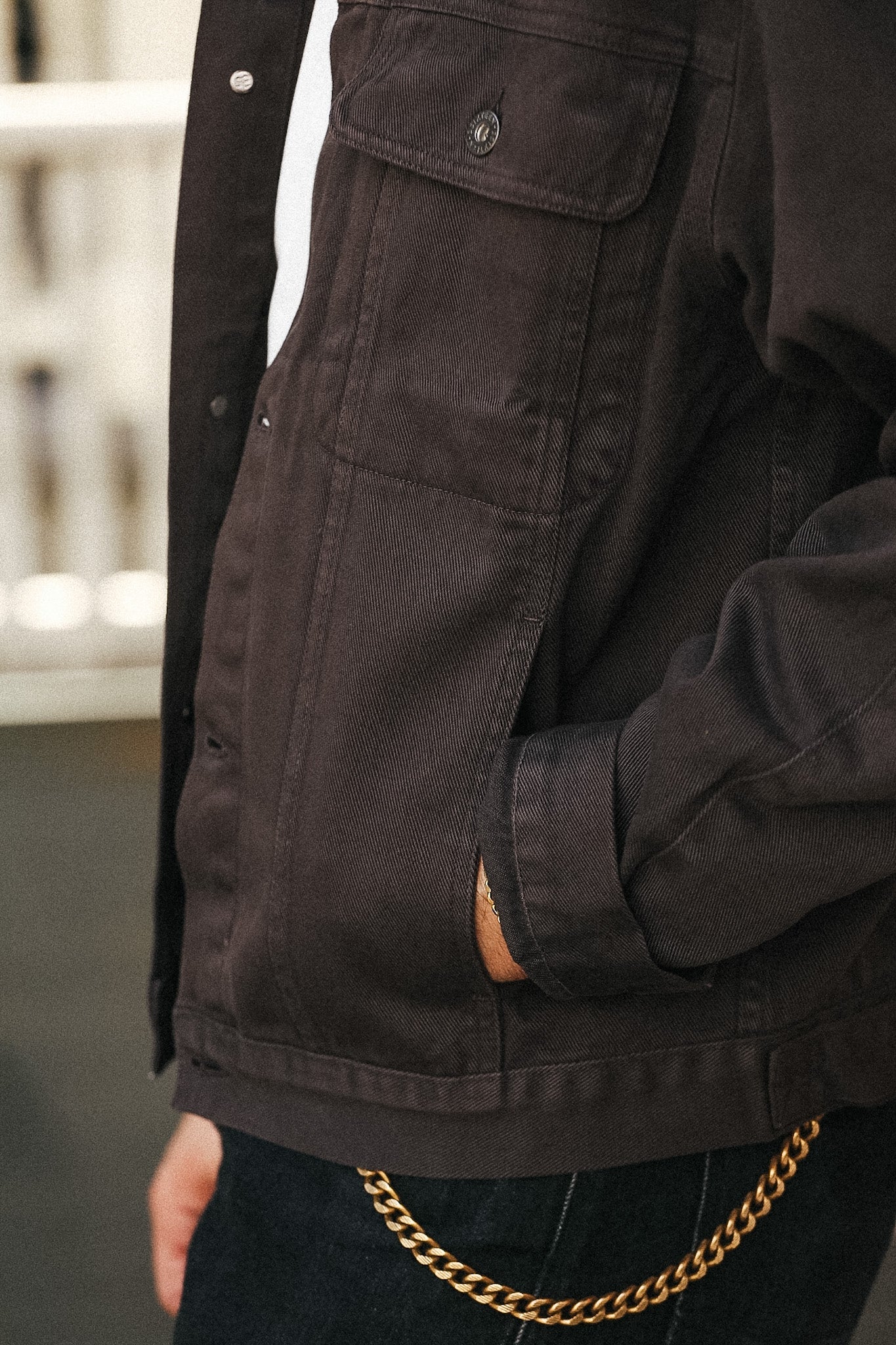 Close-up of a person wearing a dark brown canvas utility jacket with a chest pocket, layered over a white shirt and dark jeans, with a gold chain belt visible at the waist.