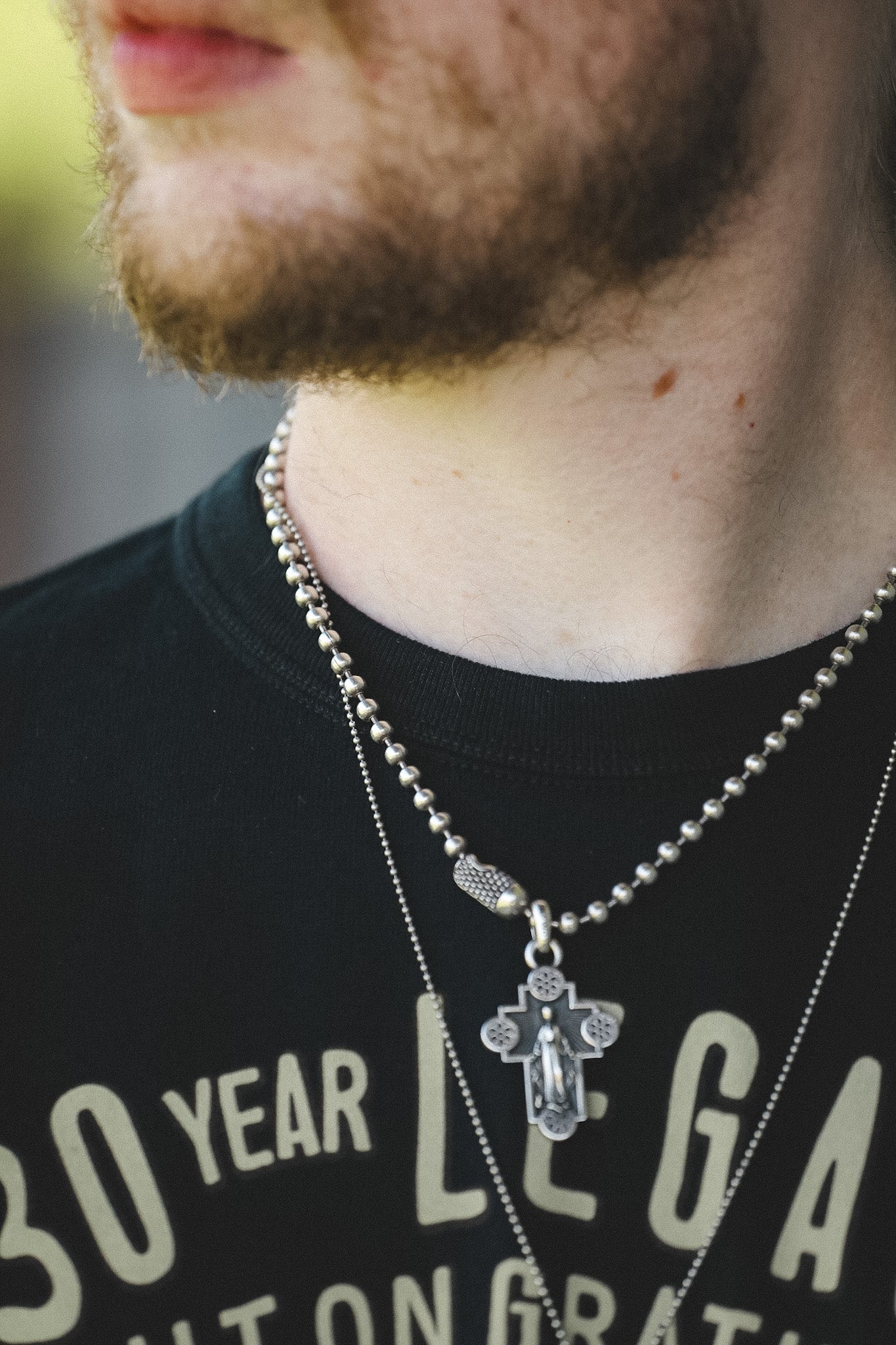 A close-up of a man's neck and lower face wearing layered silver necklaces, including a beaded chain with a cross pendant and a thinner chain with a textured charm, over a black graphic t-shirt.