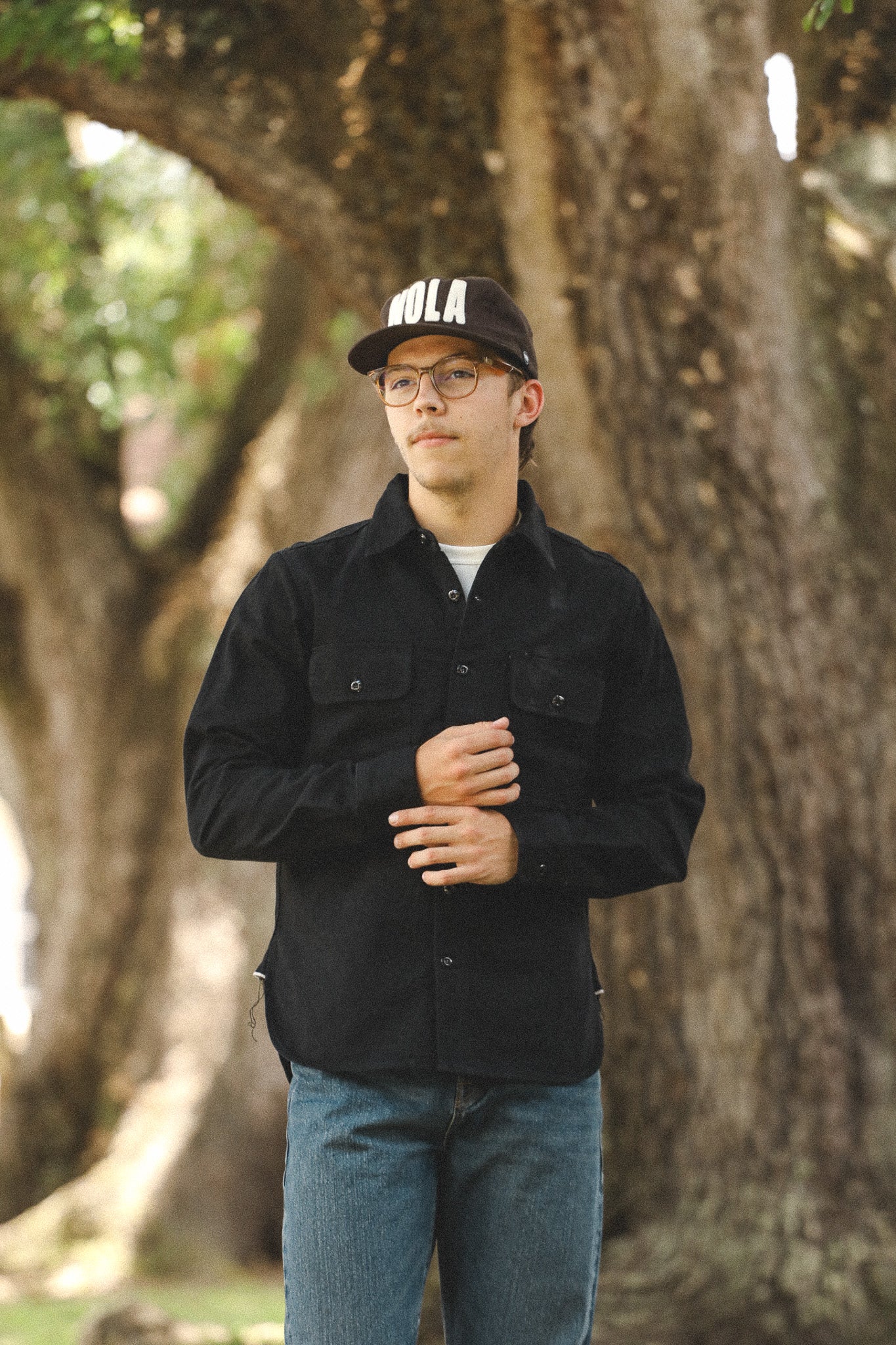 Person wearing a black jacket and cap with 'Bola' branding, standing in front of a tree.