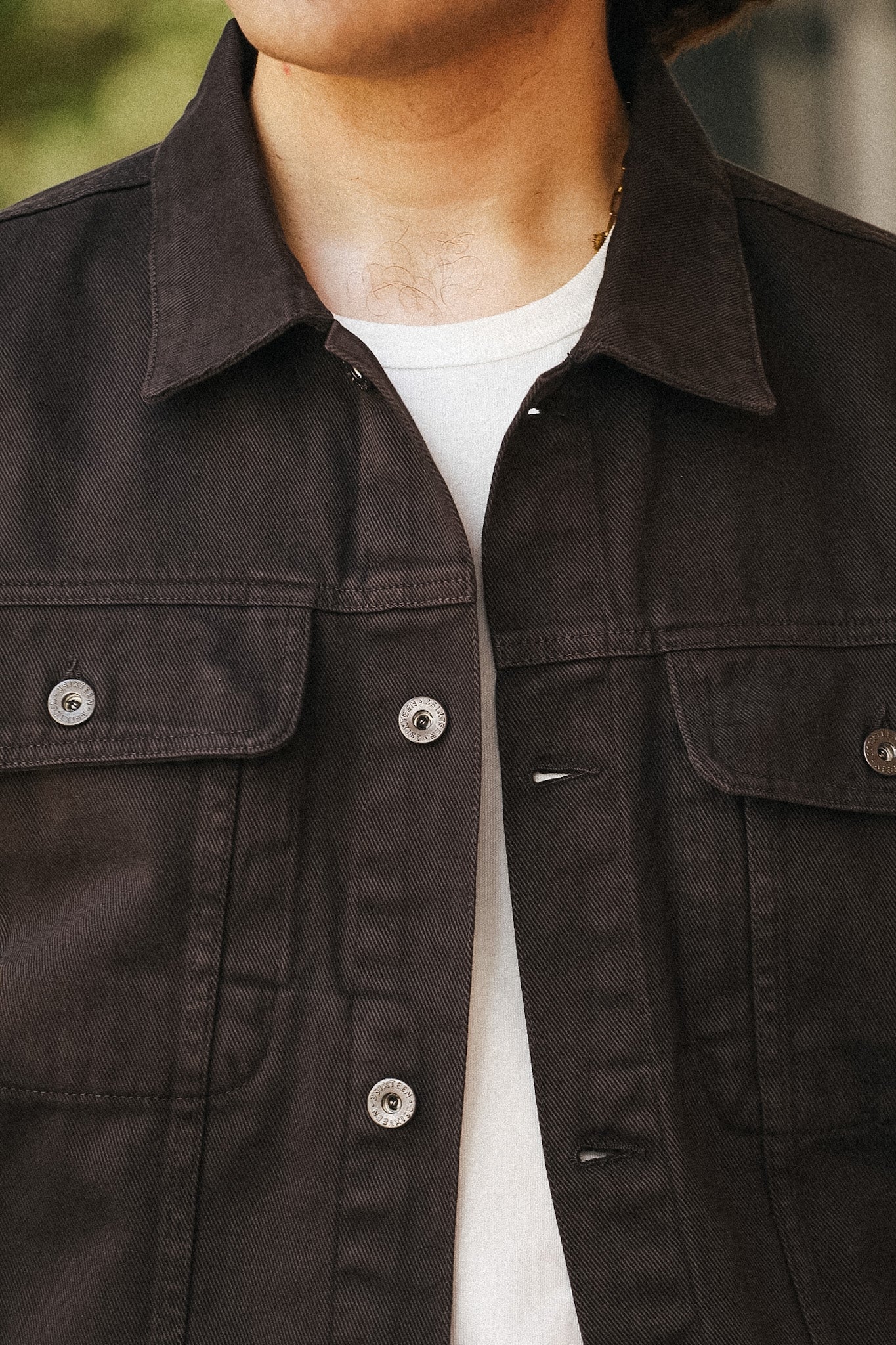 Close-up of a person wearing a dark brown or very deep charcoal button-up overshirt/jacket over a white t-shirt, with the collar, chest pockets, and buttons visible.