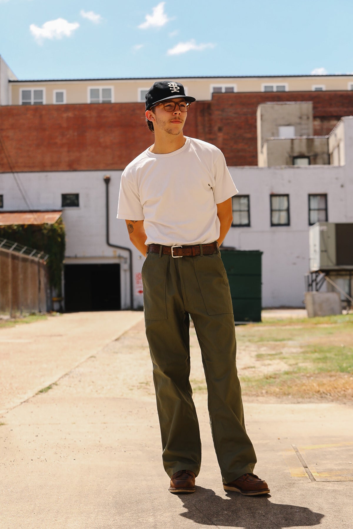 A man wearing a white t-shirt, olive green cargo pants, a brown belt, and a black baseball cap stands in an outdoor alleyway with brick buildings in the background.