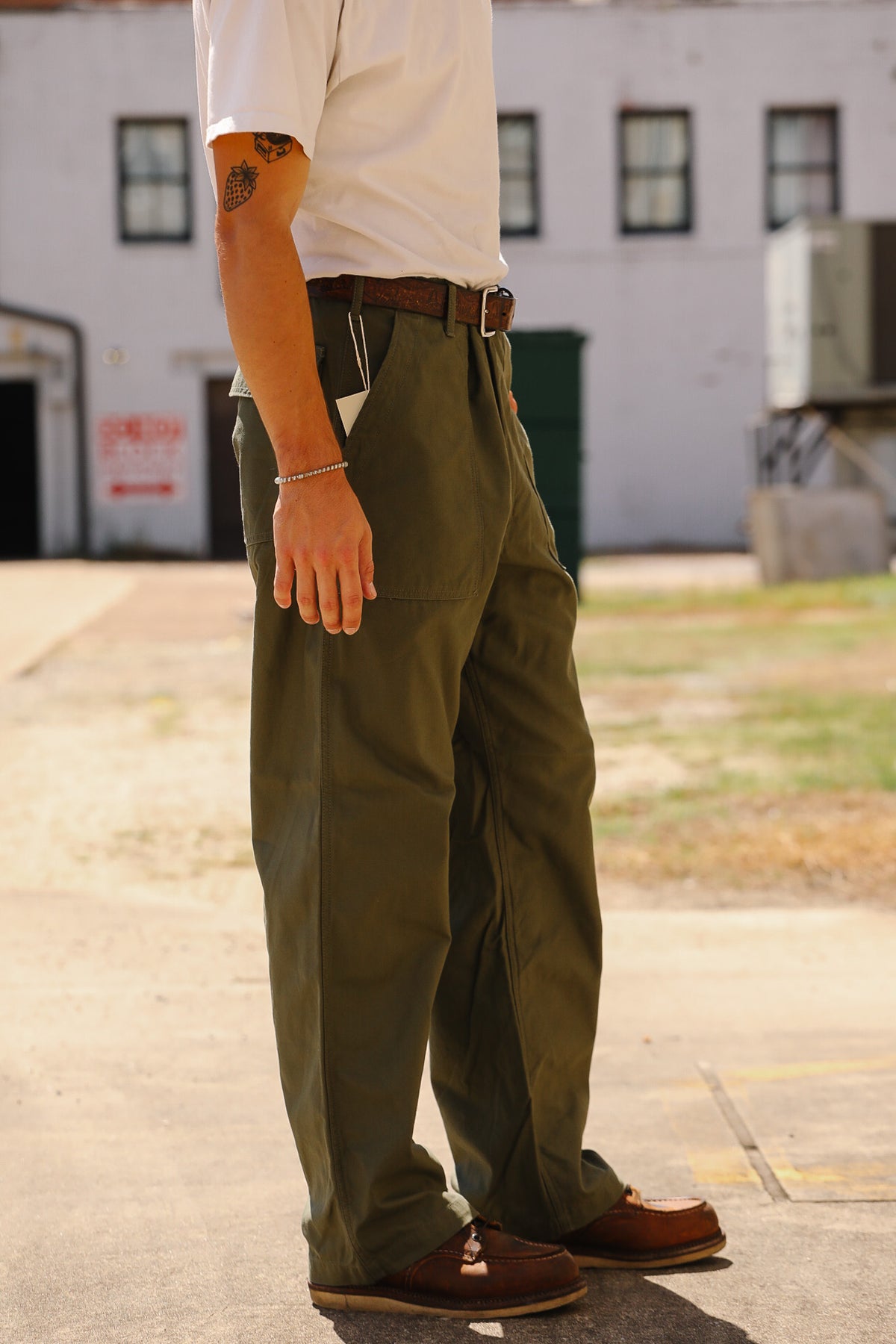 A person standing outdoors in front of a white building, wearing olive green cargo-style pants with a brown belt and brown shoes.