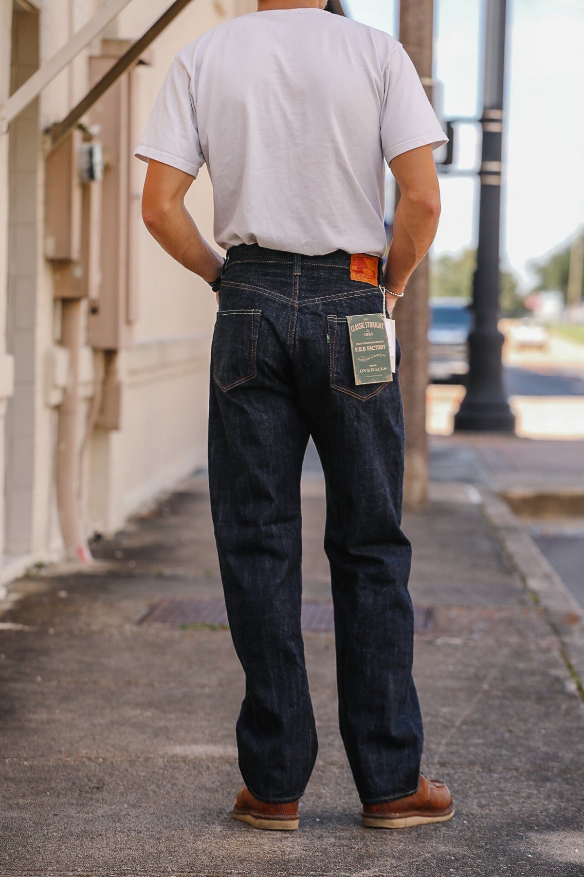 A man viewed from behind wearing dark indigo denim jeans with a green paper brand tag in the back pocket, a white t-shirt, and brown leather shoes, standing on an outdoor sidewalk.