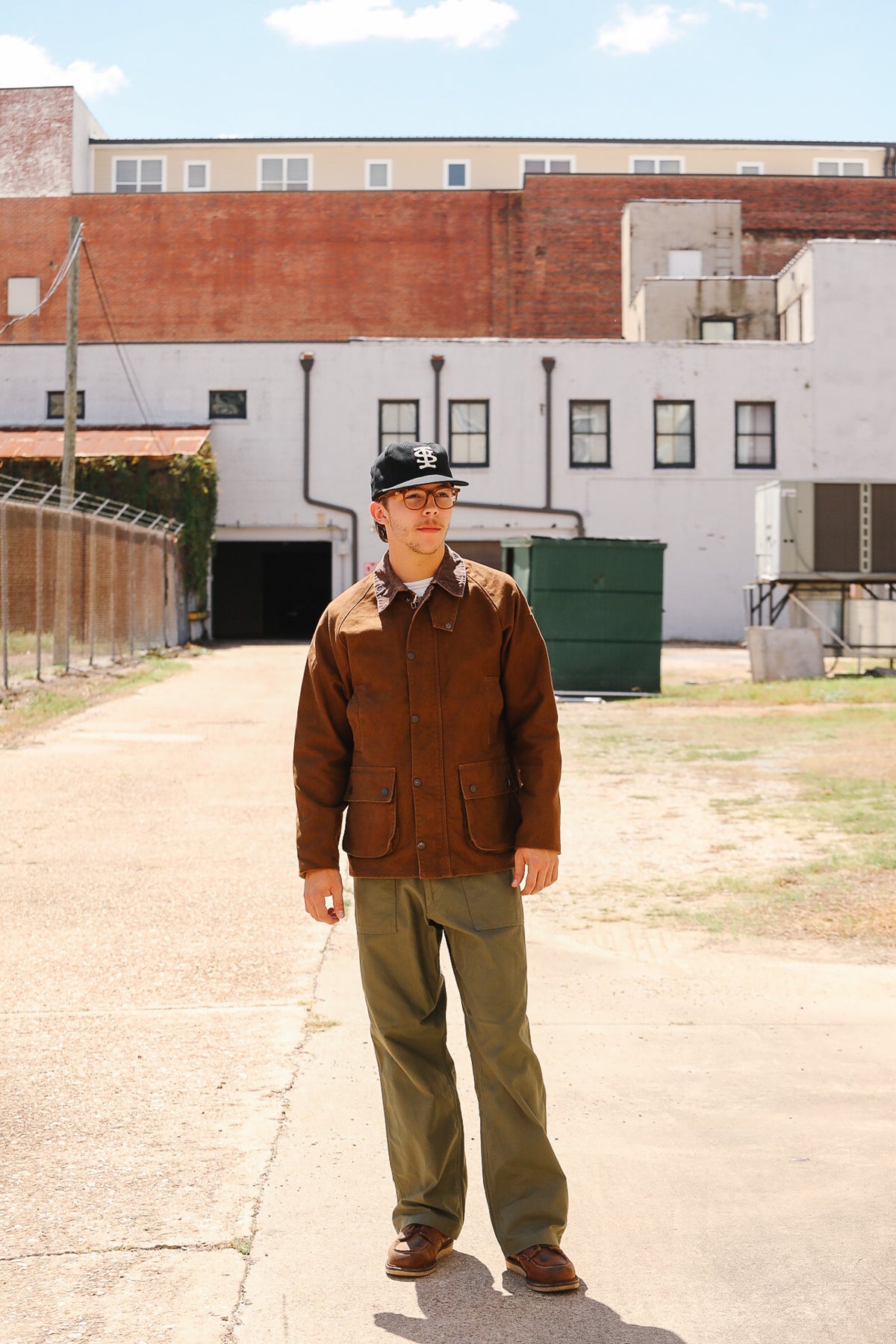 A man wearing a brown corduroy jacket, olive green pants, and a black baseball cap stands in an outdoor industrial alleyway with brick and concrete buildings in the background.
