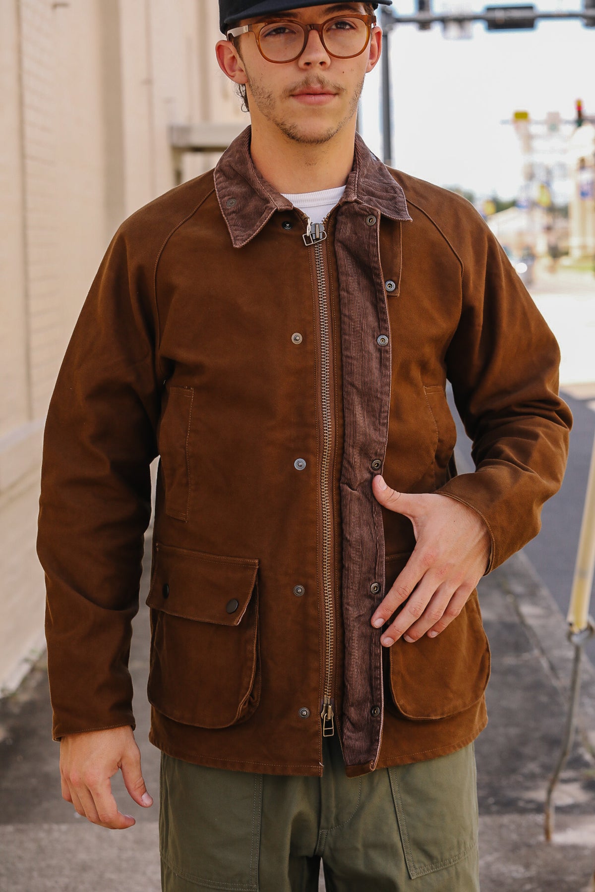 A man wearing a brown waxed canvas utility jacket with a brown corduroy collar, silver-tone metal zipper, and snap-button front, paired with olive green pants and a black cap.
