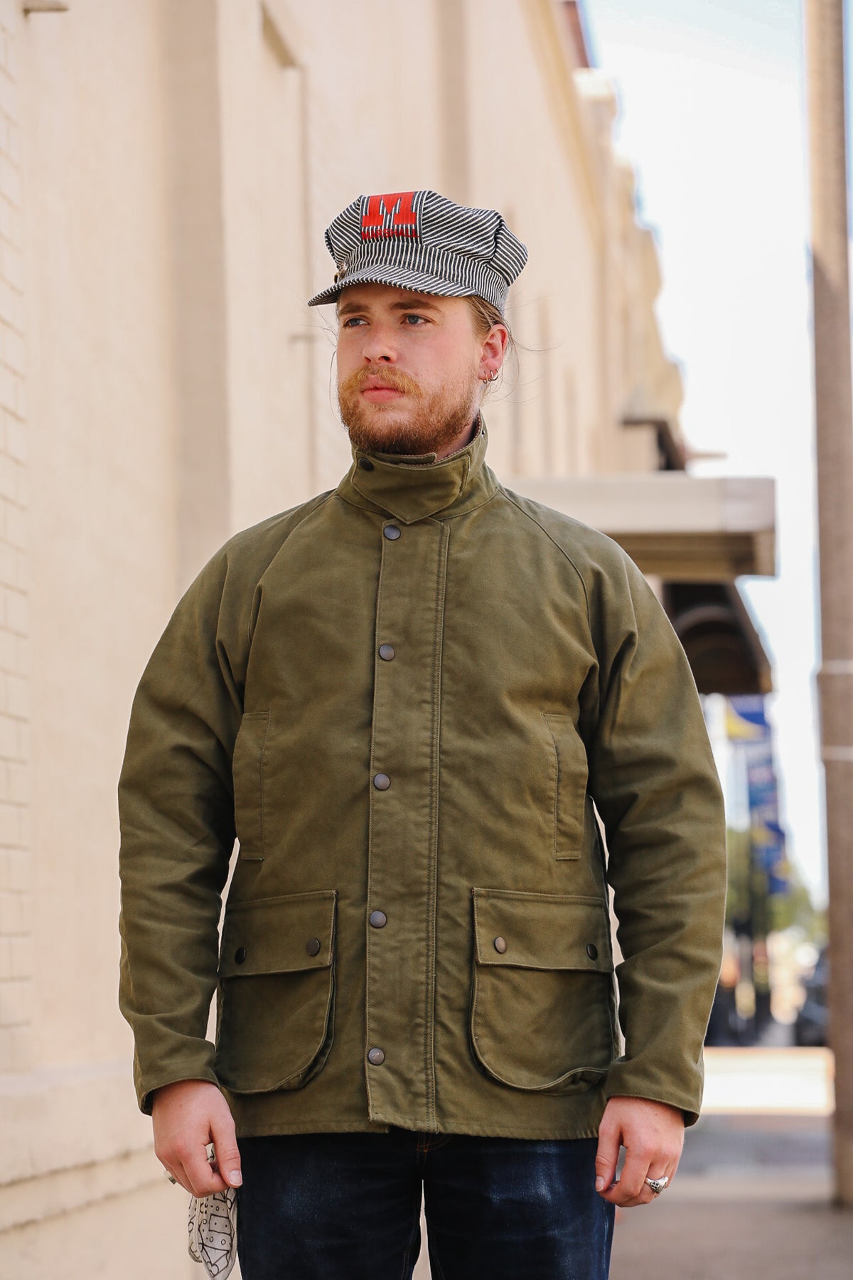 A man wearing an olive green waxed cotton utility jacket with a high collar and front pockets, paired with a gray and black striped cap, standing outdoors against a beige brick wall.