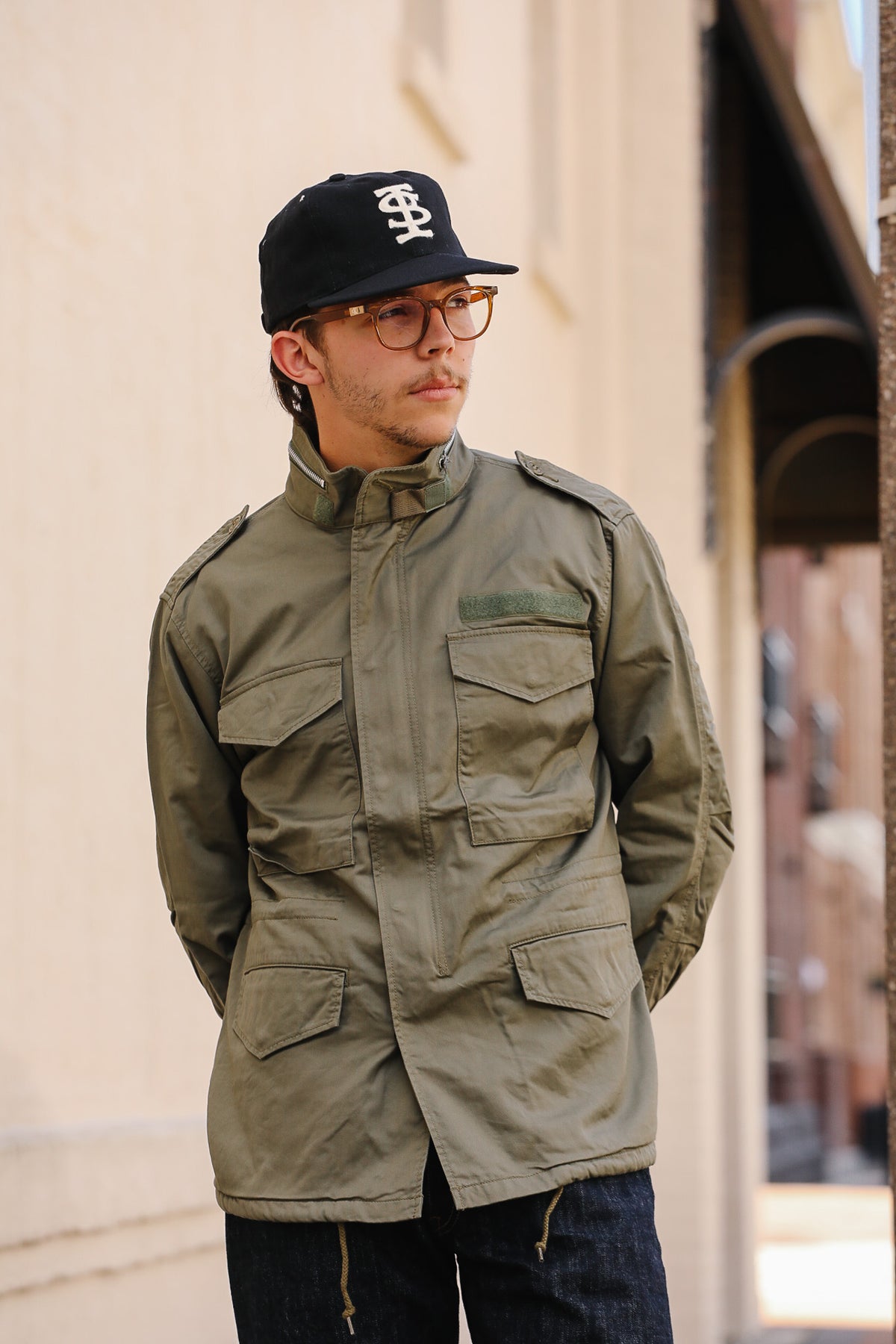 A man wearing an olive green military-style field jacket, a black baseball cap with a white monogram, and glasses, standing outdoors against a beige wall.