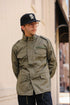 A man wearing an olive green military-style field jacket, a black baseball cap with a white monogram, and glasses, standing outdoors against a beige wall.