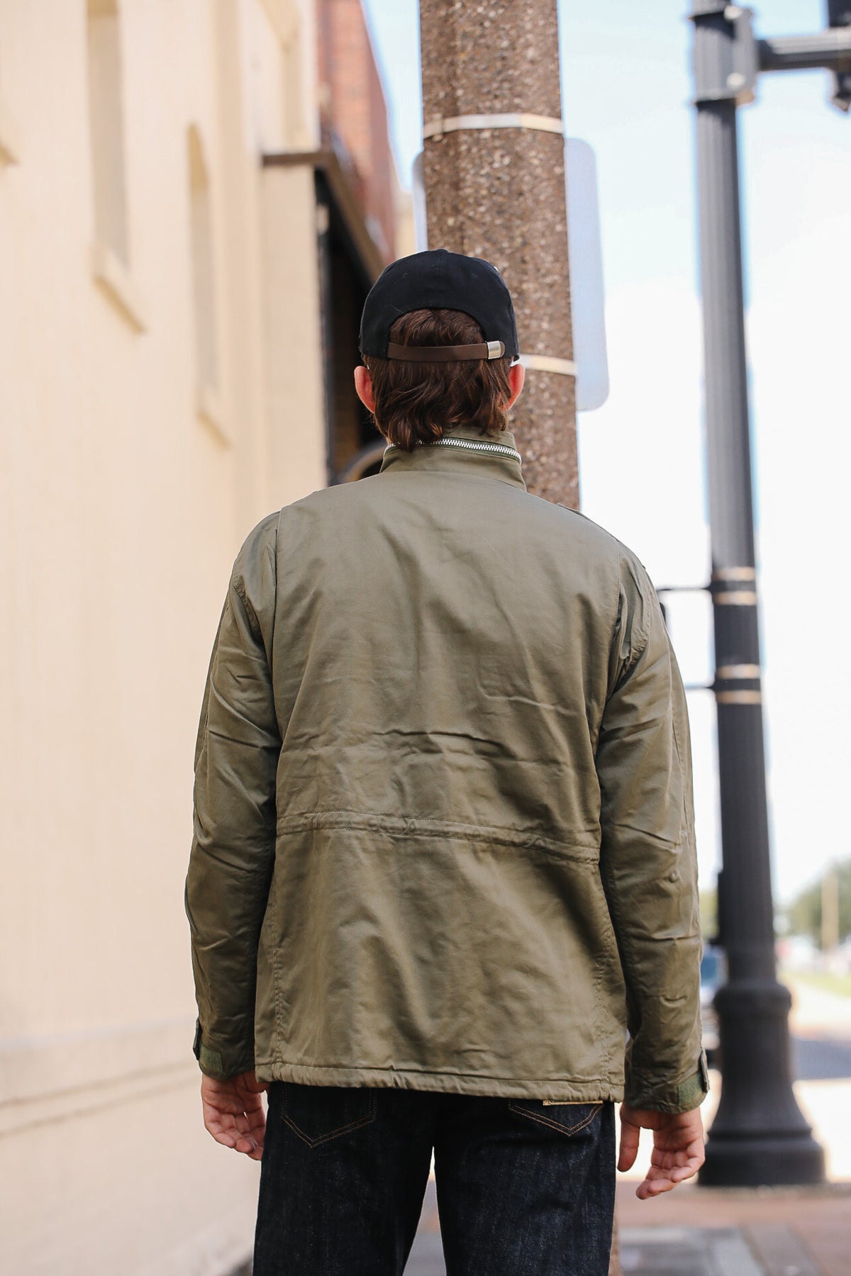 A man viewed from behind wearing an olive green lightweight jacket, a black baseball cap, and dark jeans, standing outdoors next to a beige building and a pole.