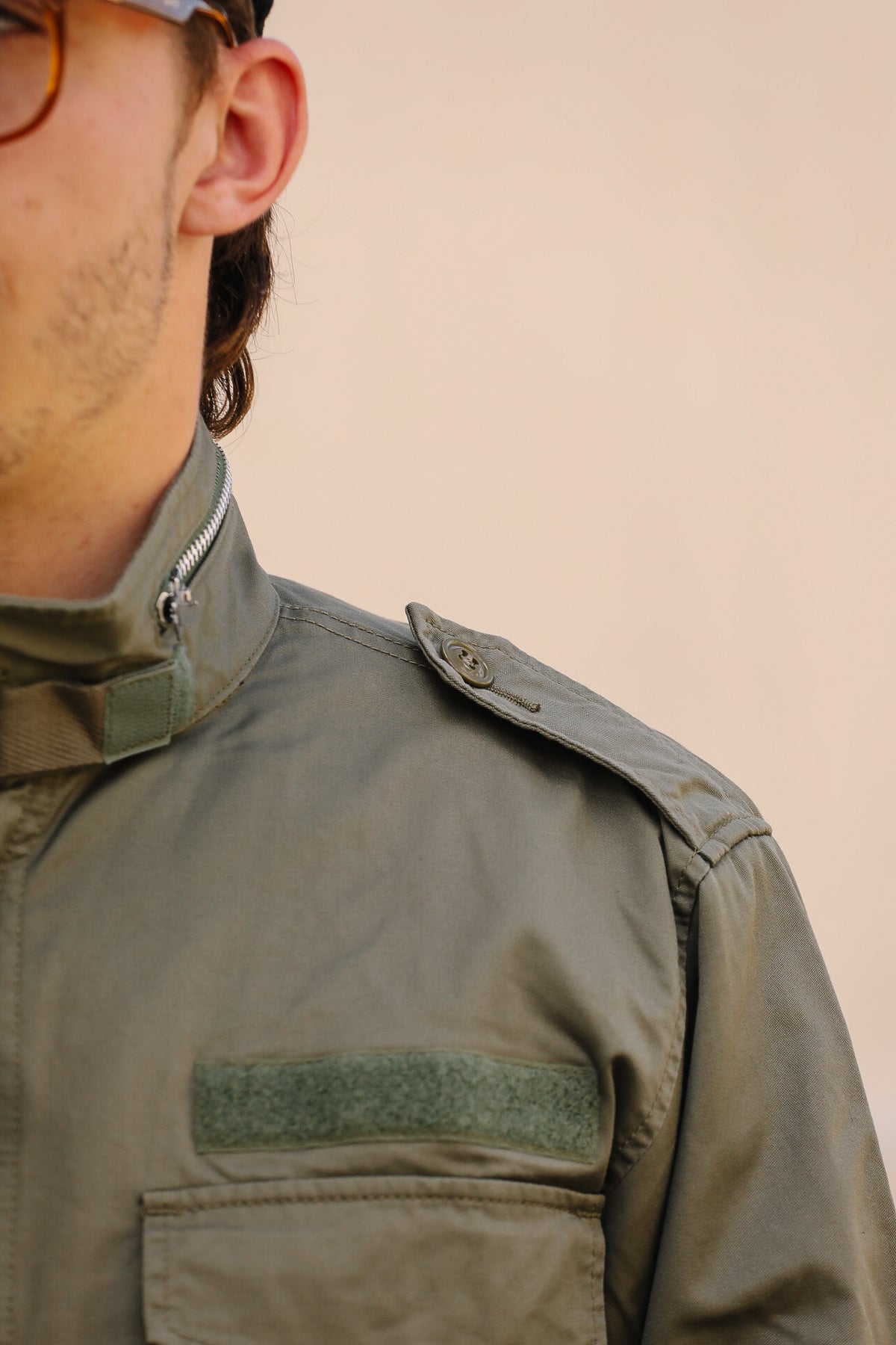 Close-up of a person wearing an olive green military-style jacket with a high collar, shoulder tab, and chest pocket against a plain beige background.