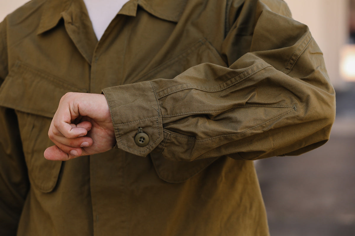 Close-up of a person wearing an olive green utility jacket, showing the buttoned cuff and chest pocket details.