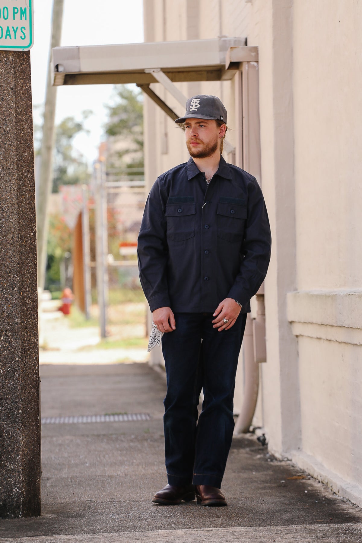 A man wearing a dark navy blue long-sleeve work shirt, dark jeans, and a baseball cap stands on a sidewalk next to a beige building.